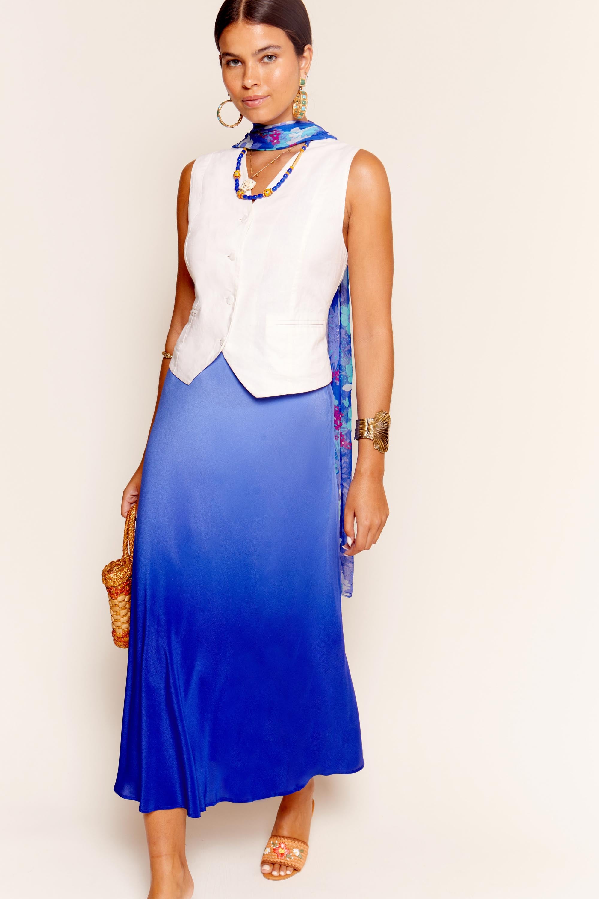 A woman wears the Norah - Cream sleeveless top over a blue maxi dress, accessorized with hoop earrings, layered necklaces, a blue patterned scarf, gold bracelet, sandals, and a small woven bag, standing against a plain background.