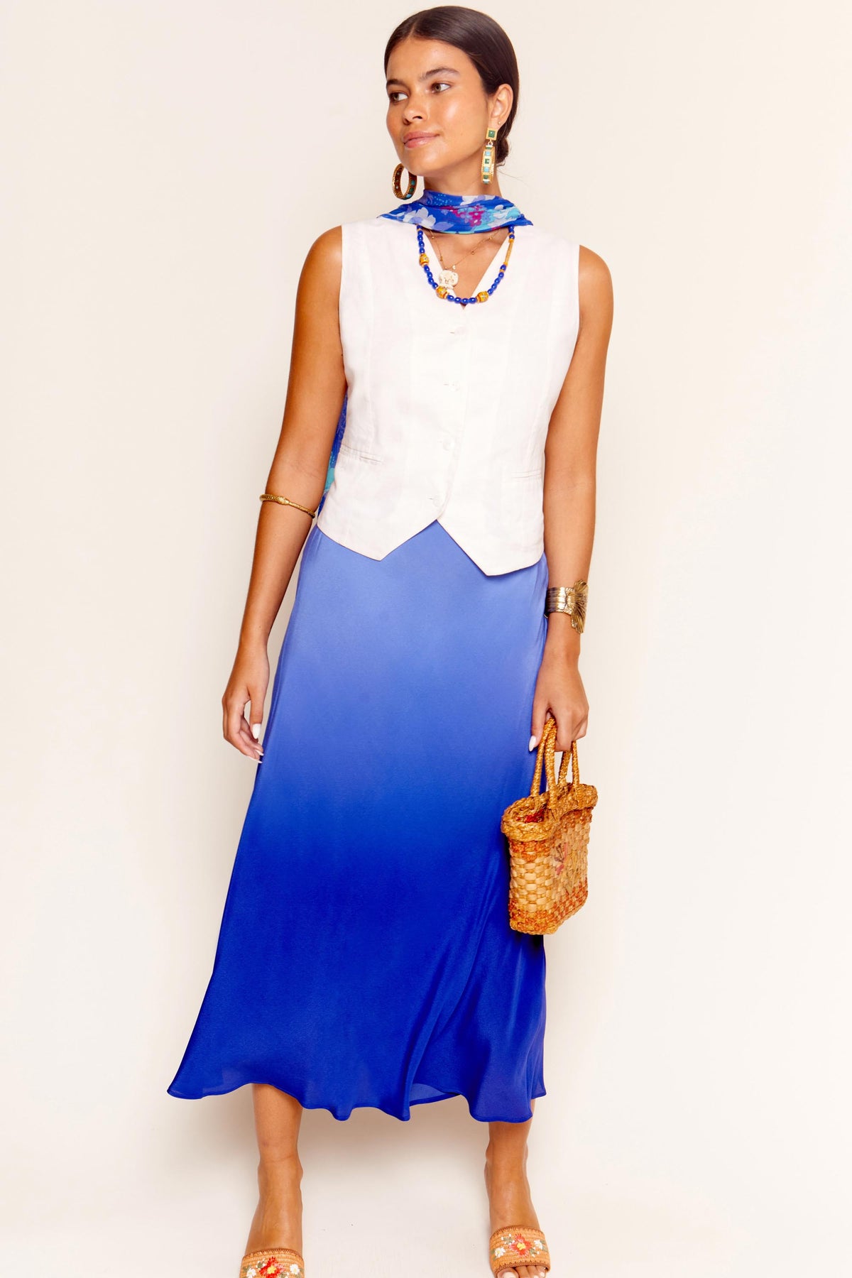 A woman stands against a plain light background wearing the Norah - Cream sleeveless top with a blue ombre maxi skirt, styled with a blue scarf, gold jewelry, statement earrings, a wicker handbag, and tan sandals.