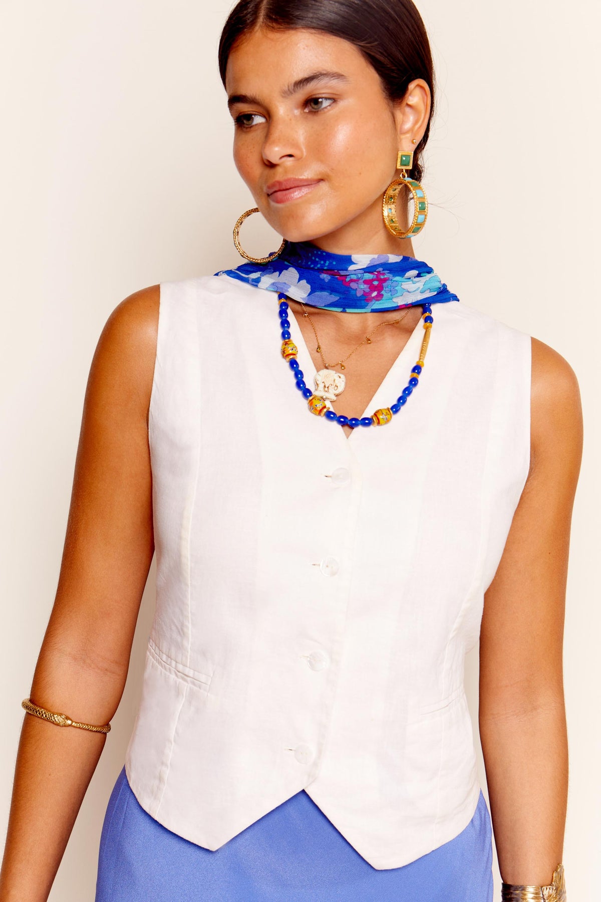 Woman wearing the Norah - Cream sleeveless top, styled with a blue floral scarf, blue beaded necklace, gold hoop earrings, and a gold bangle, looking to the side against a neutral background.