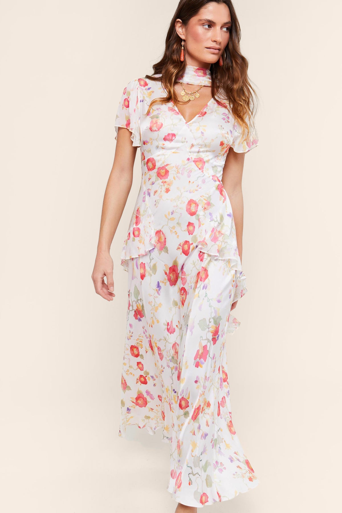 A woman wears the "evie - waterblossom ivory" dress, a long white gown with pink, red, and green floral patterns, flutter sleeves, a v-neckline, and layered ruffles—perfect for special occasions.