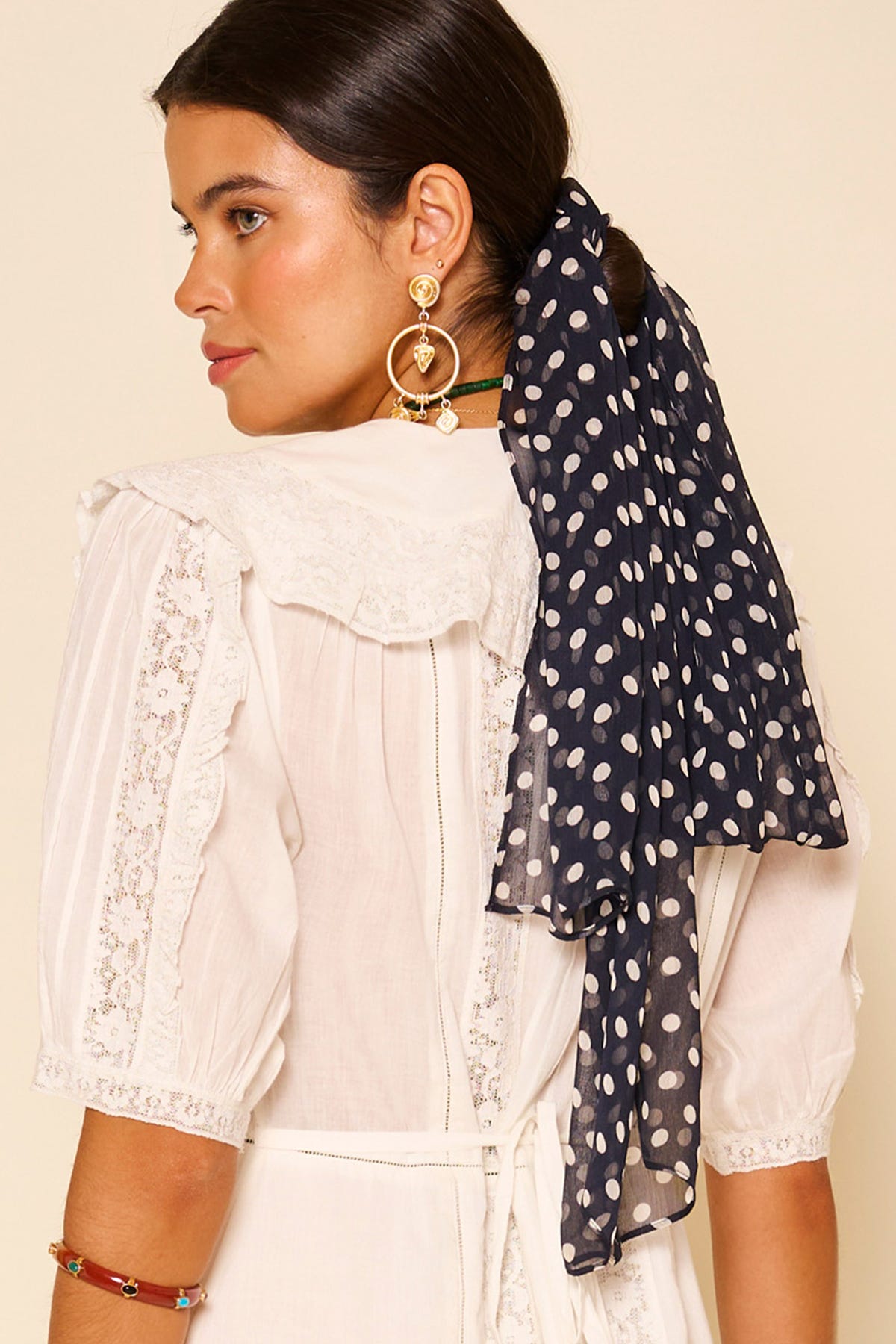 A woman with dark hair in a low ponytail, tied with the Julien - Emma Spot Deep Navy scarf, wears a white lace-trimmed dress and gold statement earrings, looking over her shoulder.