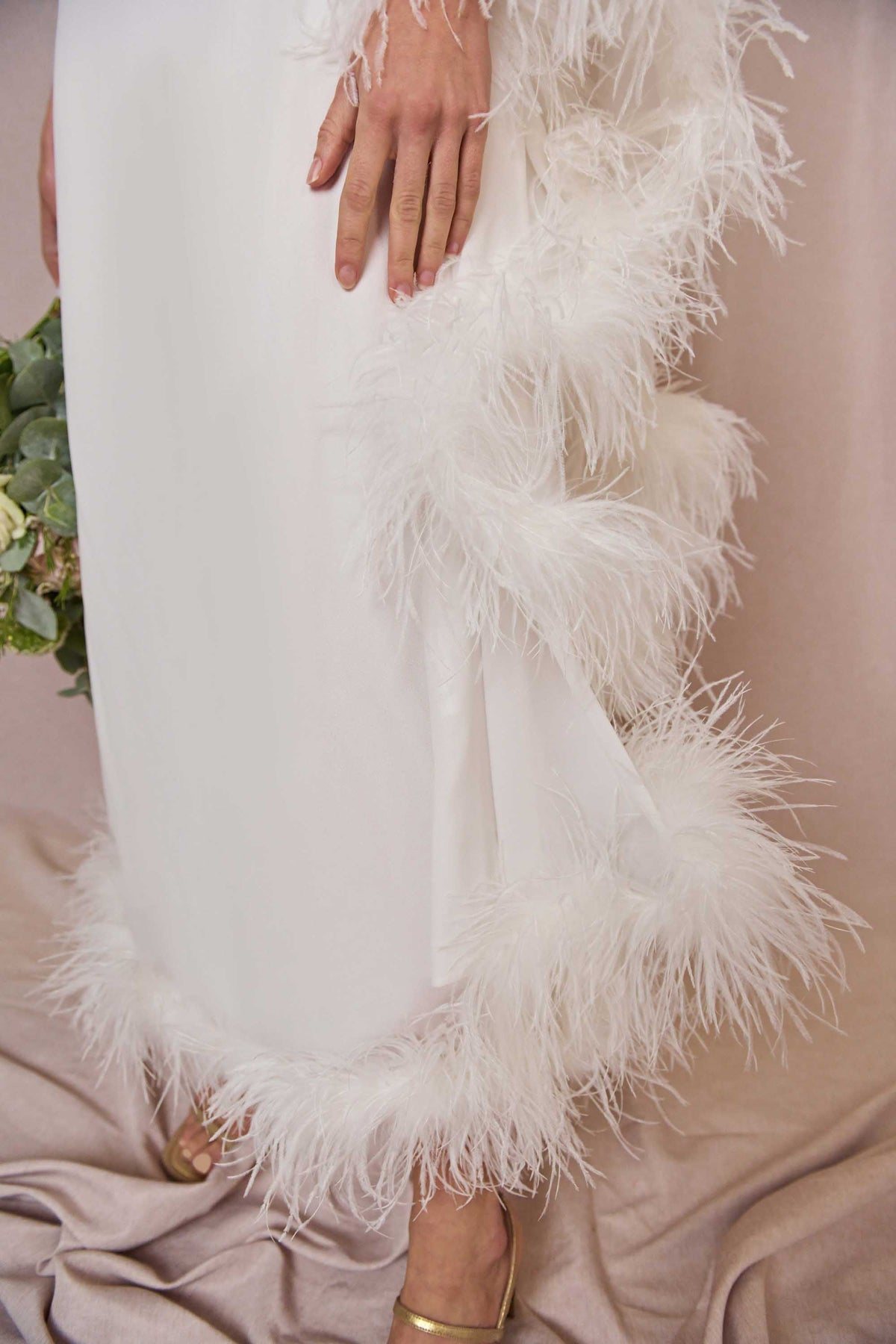 A close-up of a person wearing the Liza - Ivory Feathers dress, an ivory silk midi with white feather trim on the sleeves and hem, holding flowers and pairing it with gold open-toe shoes.