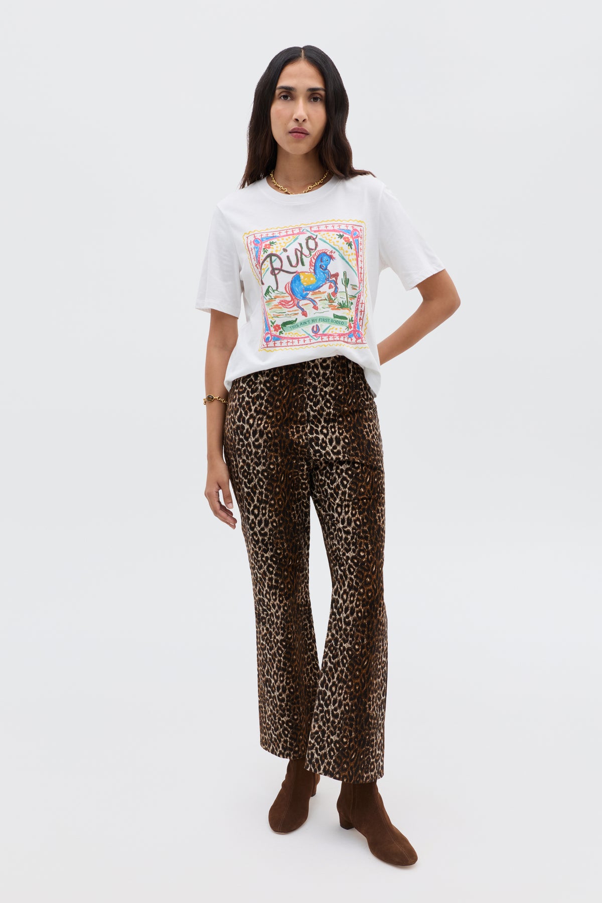 A woman stands against a plain background wearing a white printed t-shirt with colorful graphics, Olwen - Lyra Leopard Black trousers, brown ankle boots, and a gold bracelet.