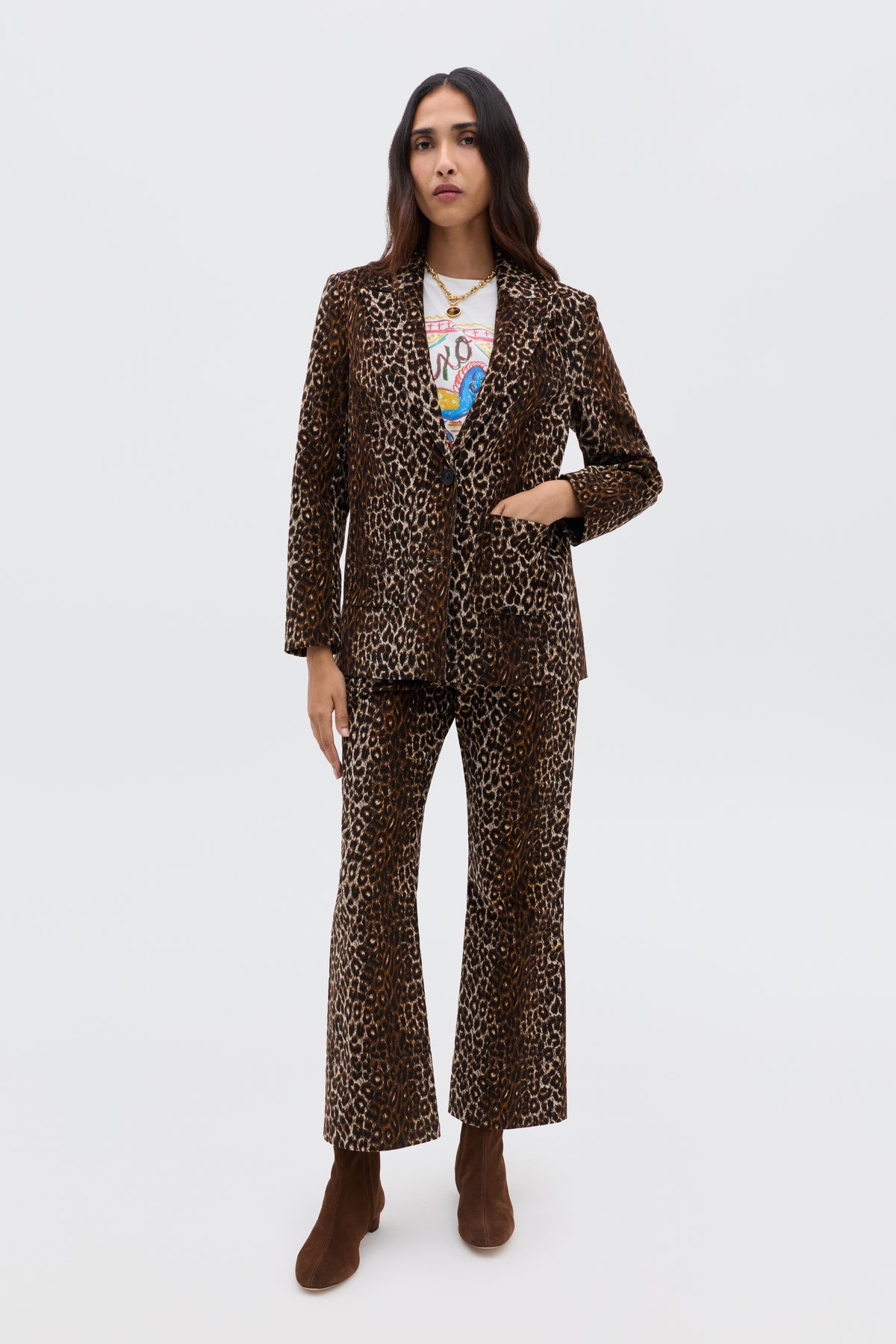A woman with long dark hair and a neutral expression stands against a plain background wearing the Olwen - Lyra Leopard Black suit, styled with vintage-inspired trousers, a white graphic tee, brown boots, and layered necklaces. One hand rests by her side.
