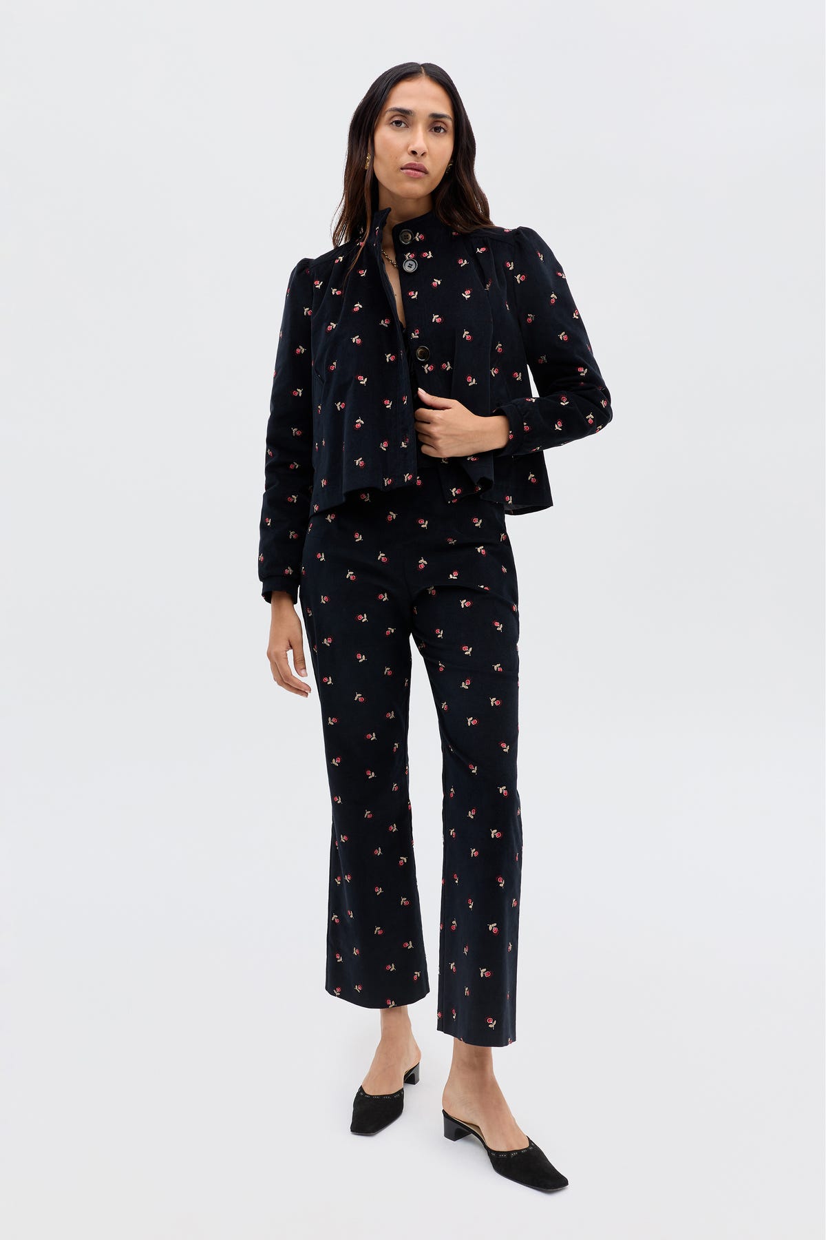 A woman stands against a plain background wearing the Olwen - Dandy Embroidery Deep Navy set, featuring vintage-inspired tailored trousers and a matching top with black floral embroidery, paired with black low-heeled shoes.