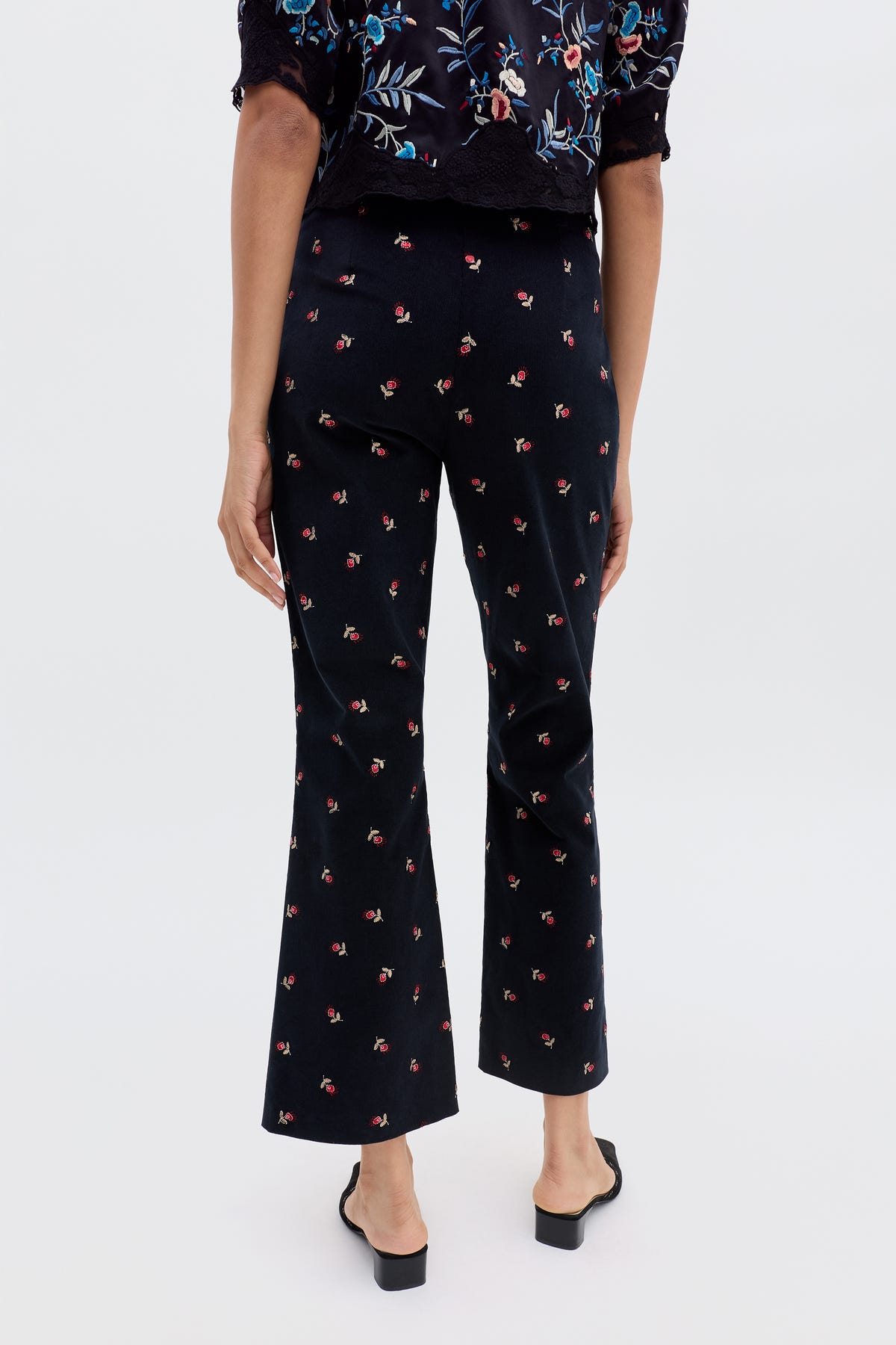 Wearing the Olwen - Dandy Embroidery Deep Navy top with blue and red floral embroidery, floral print trousers featuring a small red pattern, and black open-back heeled shoes, against a plain light background.