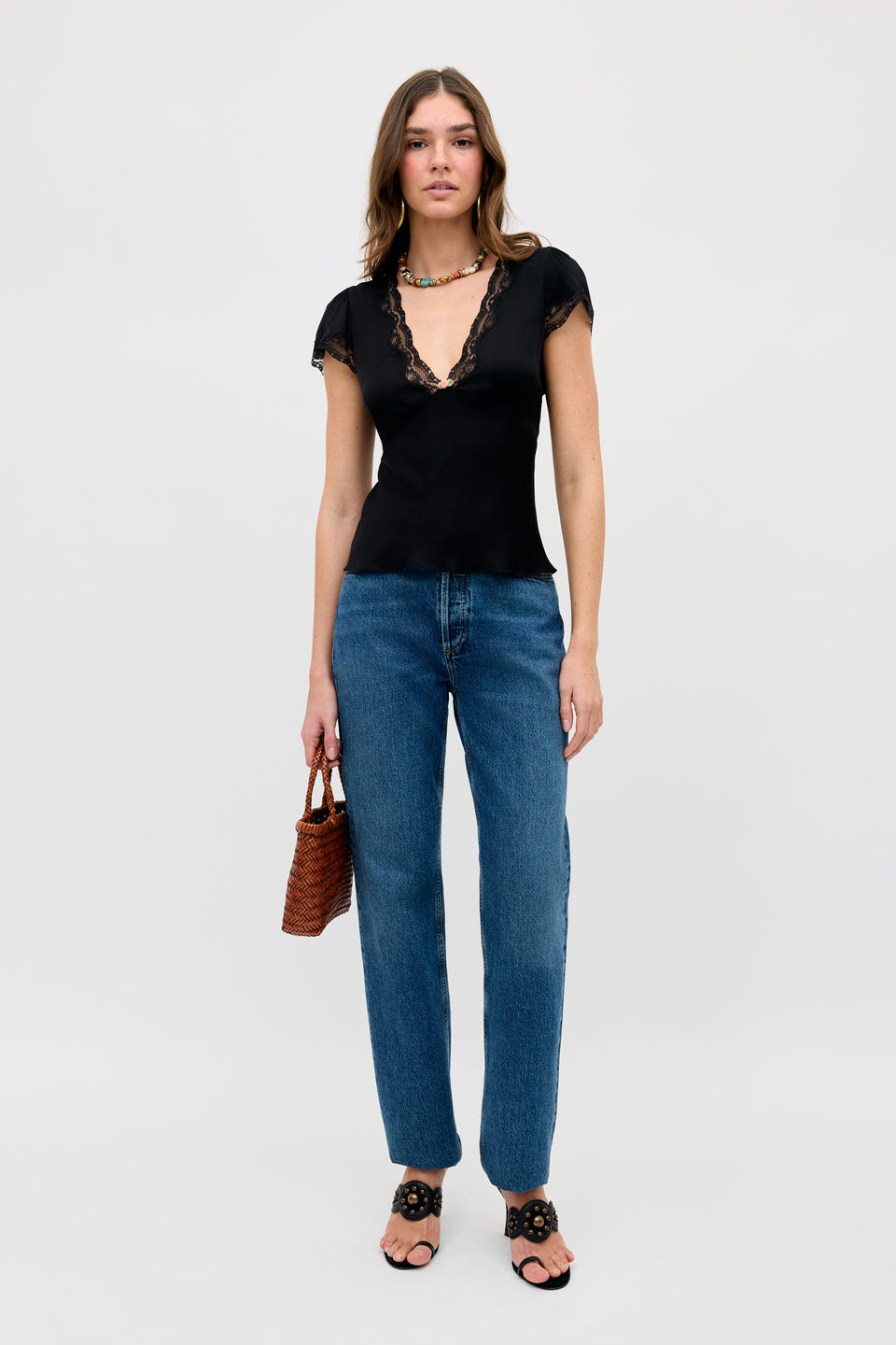 A woman stands against a plain background wearing the Karia - Black lace-trimmed top, blue jeans, black sandals, a beaded necklace, and holding a small brown woven handbag.