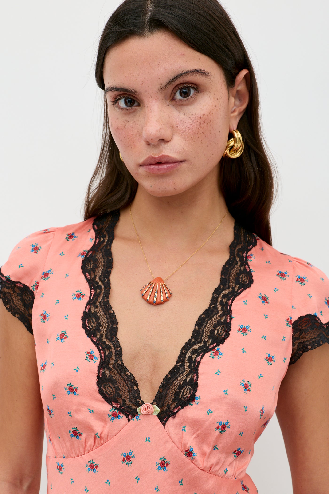 A woman with long brown hair and freckles wears the Karia - Peonies Ditsy Coral dress, paired with gold hoop earrings and a gold necklace featuring a red shell-shaped pendant, standing against a plain white background.
