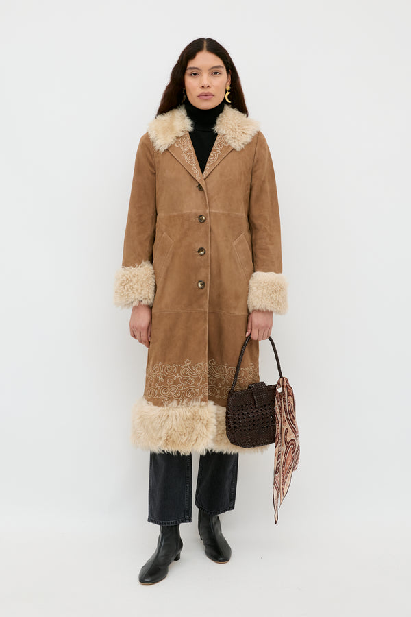 A woman stands against a white background wearing the Cienna - Camel coat, featuring cream shearling trim and an embroidered lapel, paired with a black turtleneck, black jeans, black boots, and holding a brown woven bag with a patterned scarf.