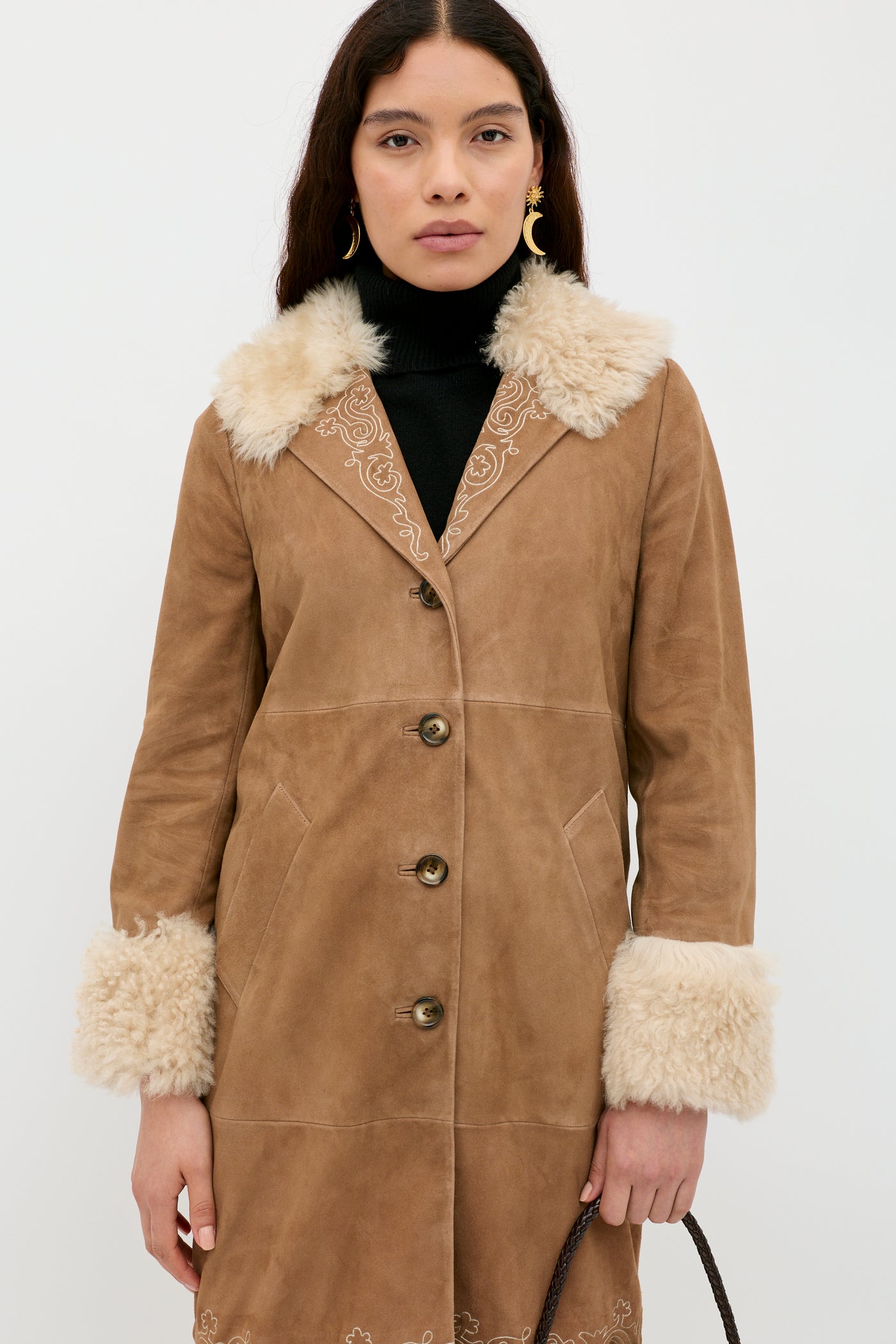 A woman with long dark hair and gold hoop earrings wears the Cienna - Camel tan suede coat with white faux fur collar and cuffs over a black turtleneck, standing against a light background.