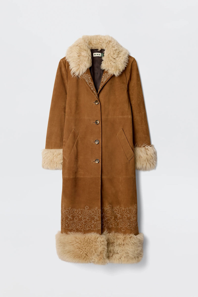 Cienna - Camel: Long brown suede coat with tan shearling collar, cuffs, and hem. Features button-down front, side pockets, embroidered lapel details, subtle decorative embroidery near the hem, and soft brown lining.
