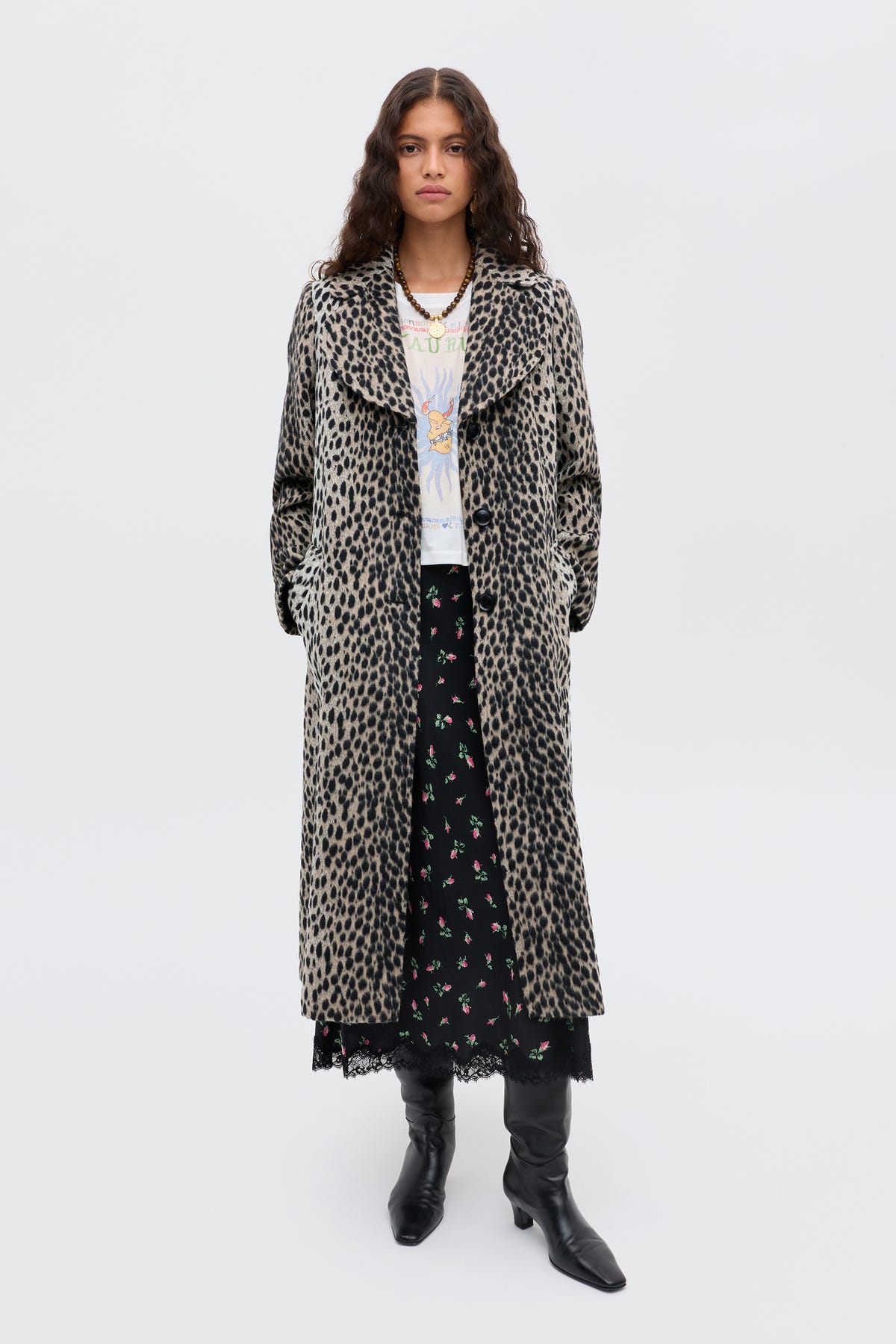 A woman with long, wavy hair and a neutral expression stands against a plain background wearing the Rosemary - Antelope Spot Jacquard Black coat over a white graphic tee, black floral skirt, and black knee-high boots.