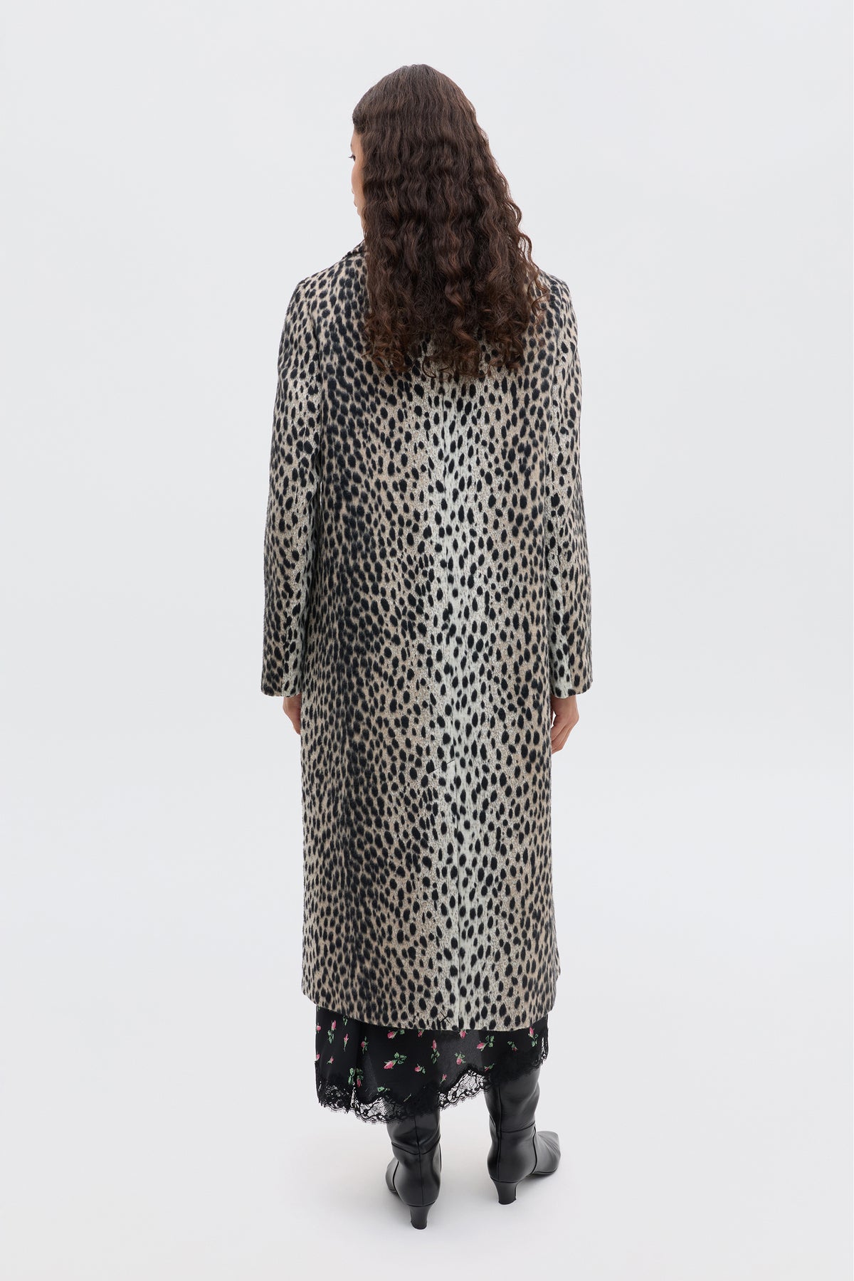A person with long curly hair, seen from behind, wears the Rosemary - Antelope Spot Jacquard Black coat over a floral black skirt and black knee-high boots, standing against a plain white background.