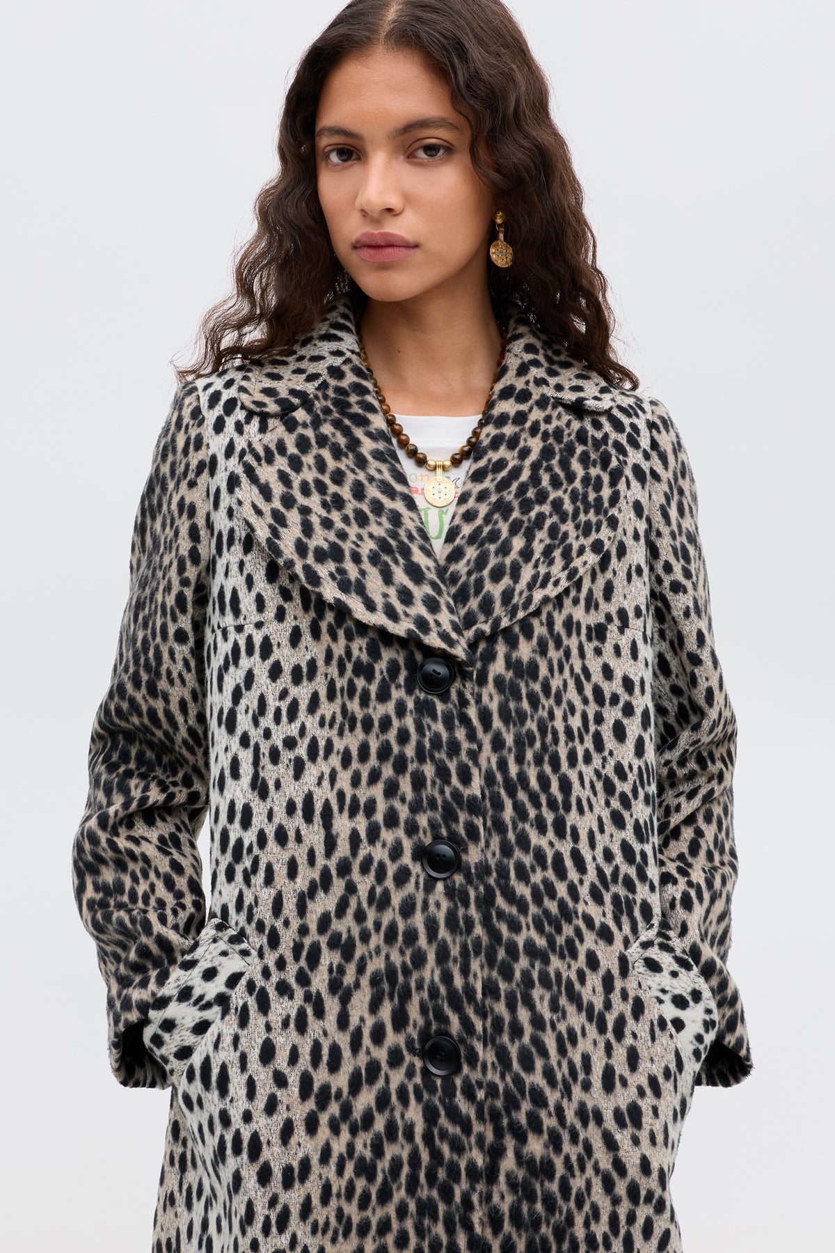 A woman with long, wavy brown hair and a neutral expression wears the Rosemary - Antelope Spot Jacquard Black coat, featuring large black buttons and pockets. She stands against a plain light background.