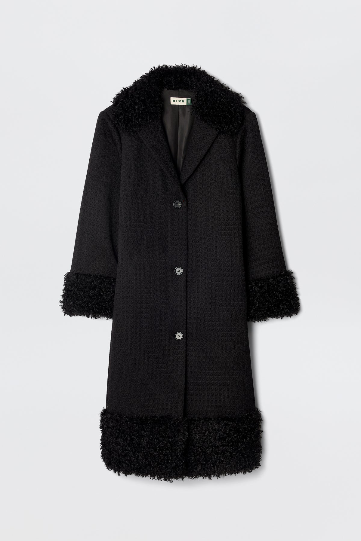 The Diara - Black is a long, black 70s-style coat with three buttons and fluffy faux-fur shearling trim on the collar, cuffs, and hem, shown against a plain white background.