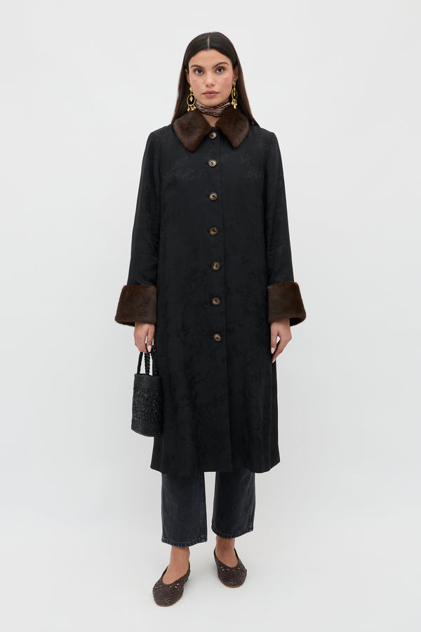 A woman stands against a plain background, wearing the Jonah - Black coat with brown fur collar and cuffs, dark pants, brown shoes, gold earrings, and holding a small black handbag. She looks directly at the camera.