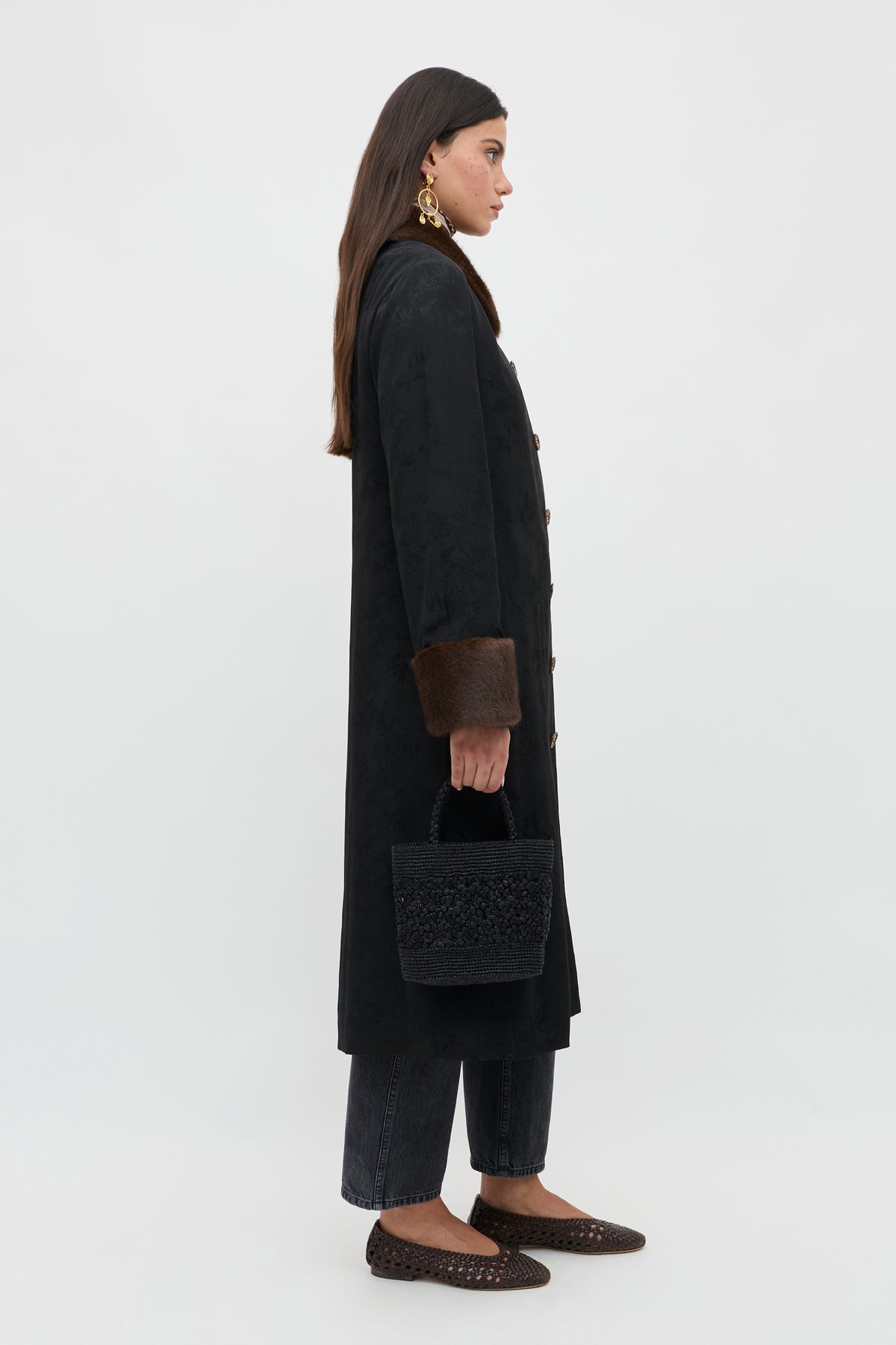 A woman stands in profile against a plain background, wearing a long black coat with brown fur cuffs, dark pants, brown shoes, gold earrings, and holding the Jonah - Black textured handbag.