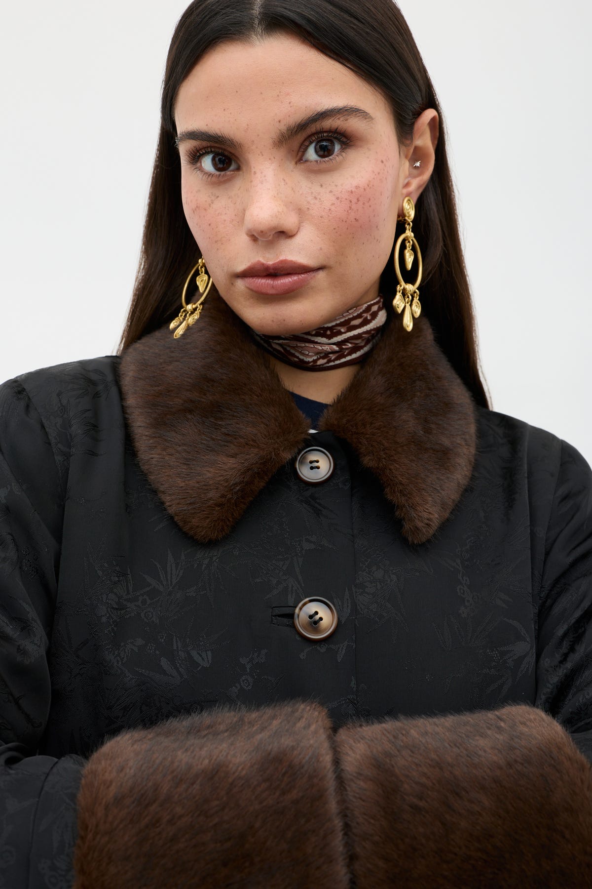 A woman with long dark hair and freckles wears the Jonah - Black coat featuring a brown fur collar and cuffs, gold earrings, and a patterned scarf, standing against a plain light background.