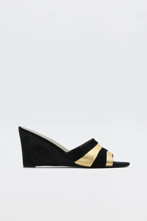 The Alira - Gold is a '70s-inspired black and gold wedge sandal with an open toe and wide crisscross straps, featuring bold gold accents. Suede-like wedge heel and straps contrast against a plain white background.