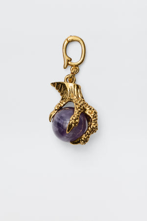 The Ianna - Gold charm features a gold-plated bird's claw gripping an Amethyst gemstone, complete with a top loop for easy attachment—ideal for vintage-inspired jewelry lovers. Photographed on a plain white background.