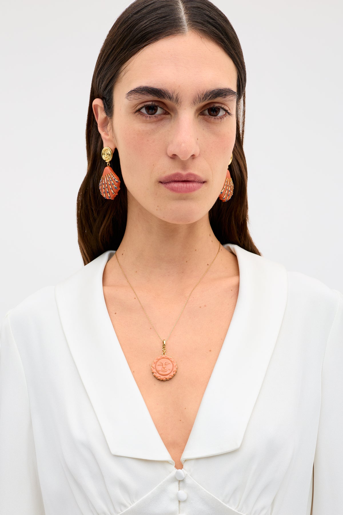 A woman with straight dark hair wears a white blouse, orange and gold earrings, and the Lillias - Orange necklace featuring a round pendant with a smiling sun face, set against a plain light background.