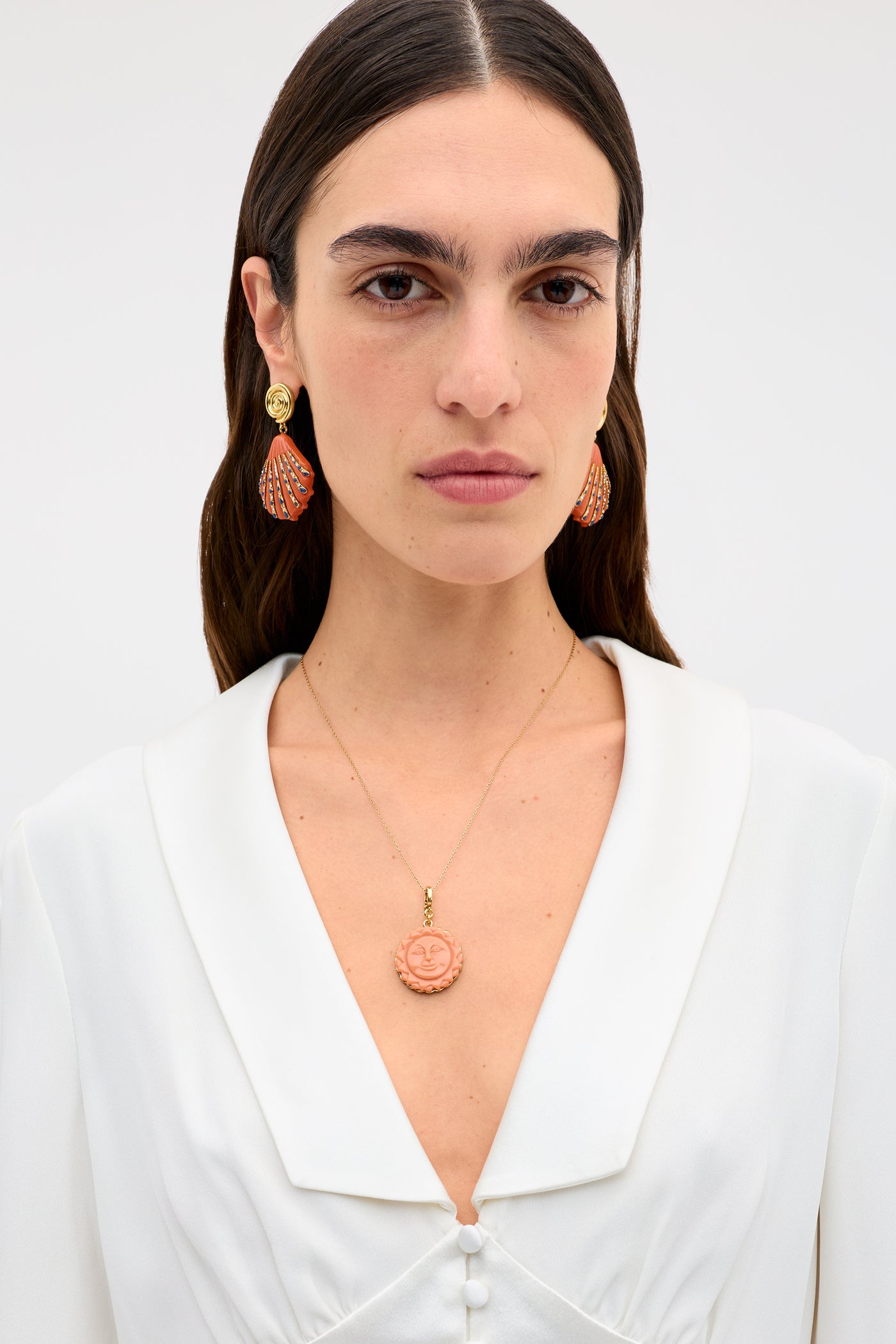 A woman with straight dark hair wears a white blouse, orange and gold earrings, and the Lillias - Orange necklace featuring a round pendant with a smiling sun face, set against a plain light background.
