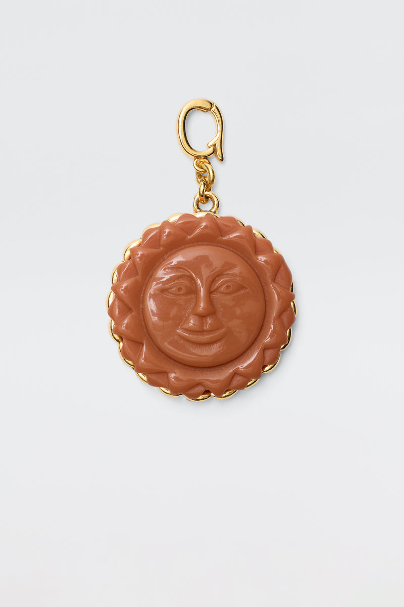 The Lillias - Orange is a round vintage charm pendant featuring a carved orange sun with a smiling face, surrounded by a gold scalloped border and attached to a gold clasp, displayed on a plain white background.