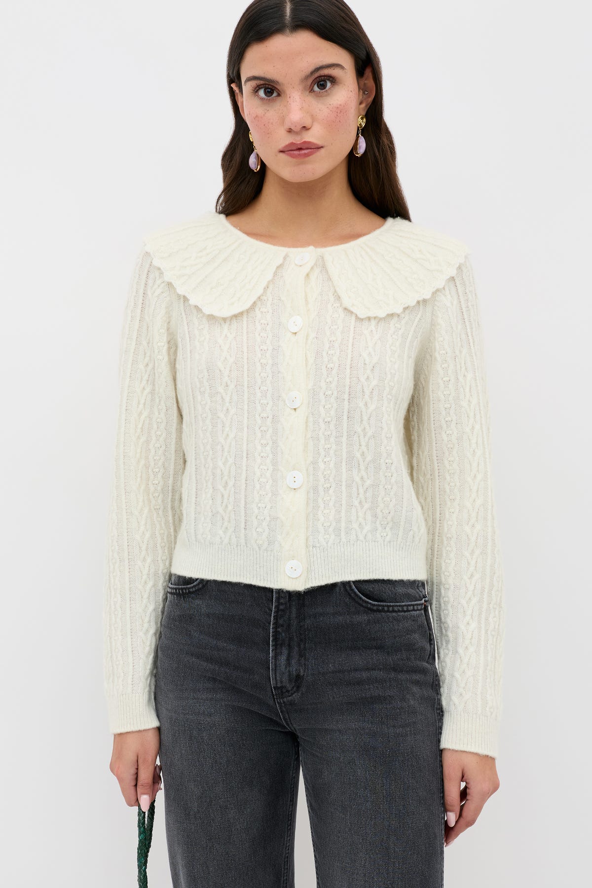 A woman stands against a plain white background wearing the Jelena - Cream button-up cable knit cardigan, featuring a large, textured collar, paired with dark gray jeans.