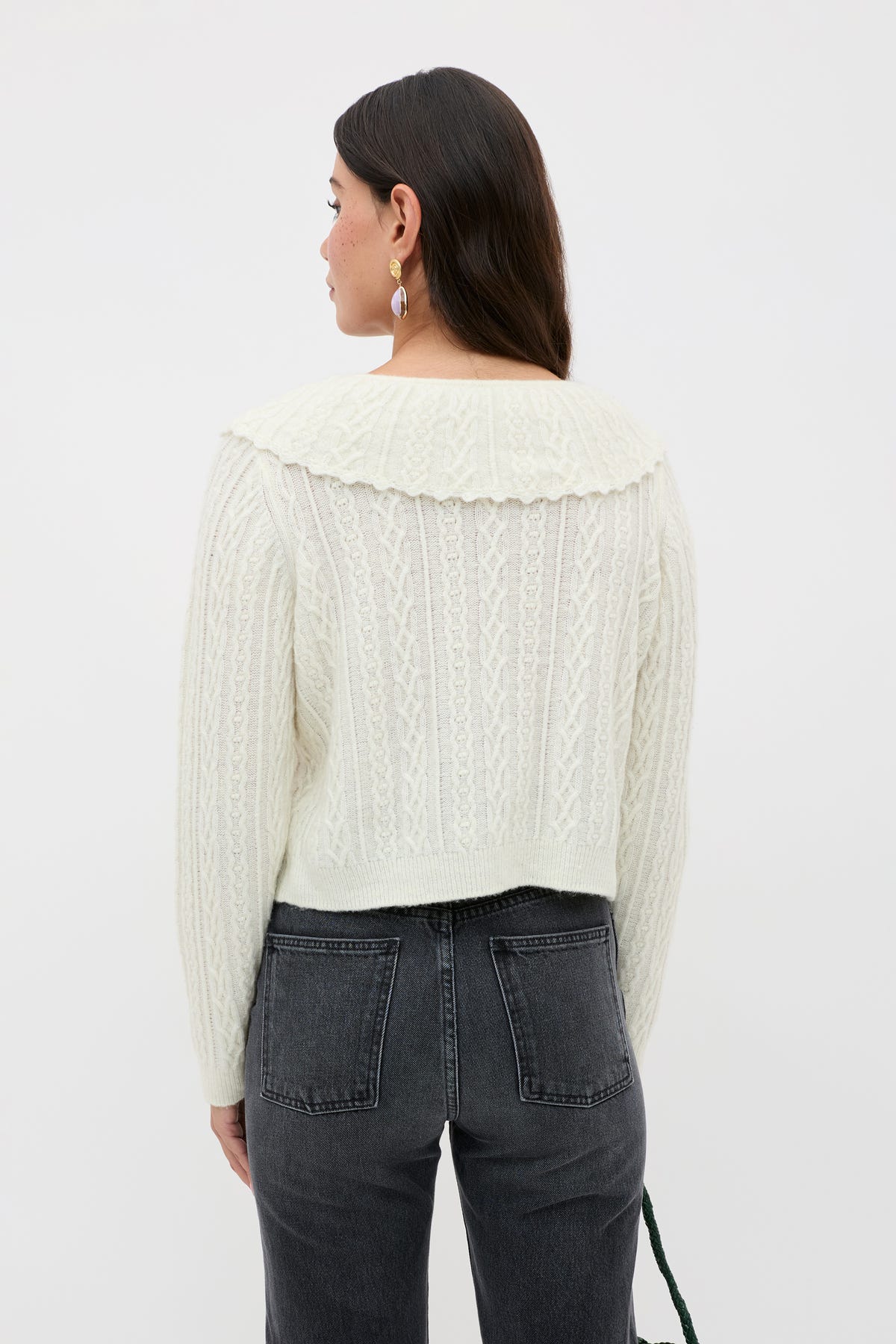 A person wearing the Jelena - Cream cable knit cardigan, featuring a wide collar, paired with dark high-waisted jeans stands facing away against a plain white background.