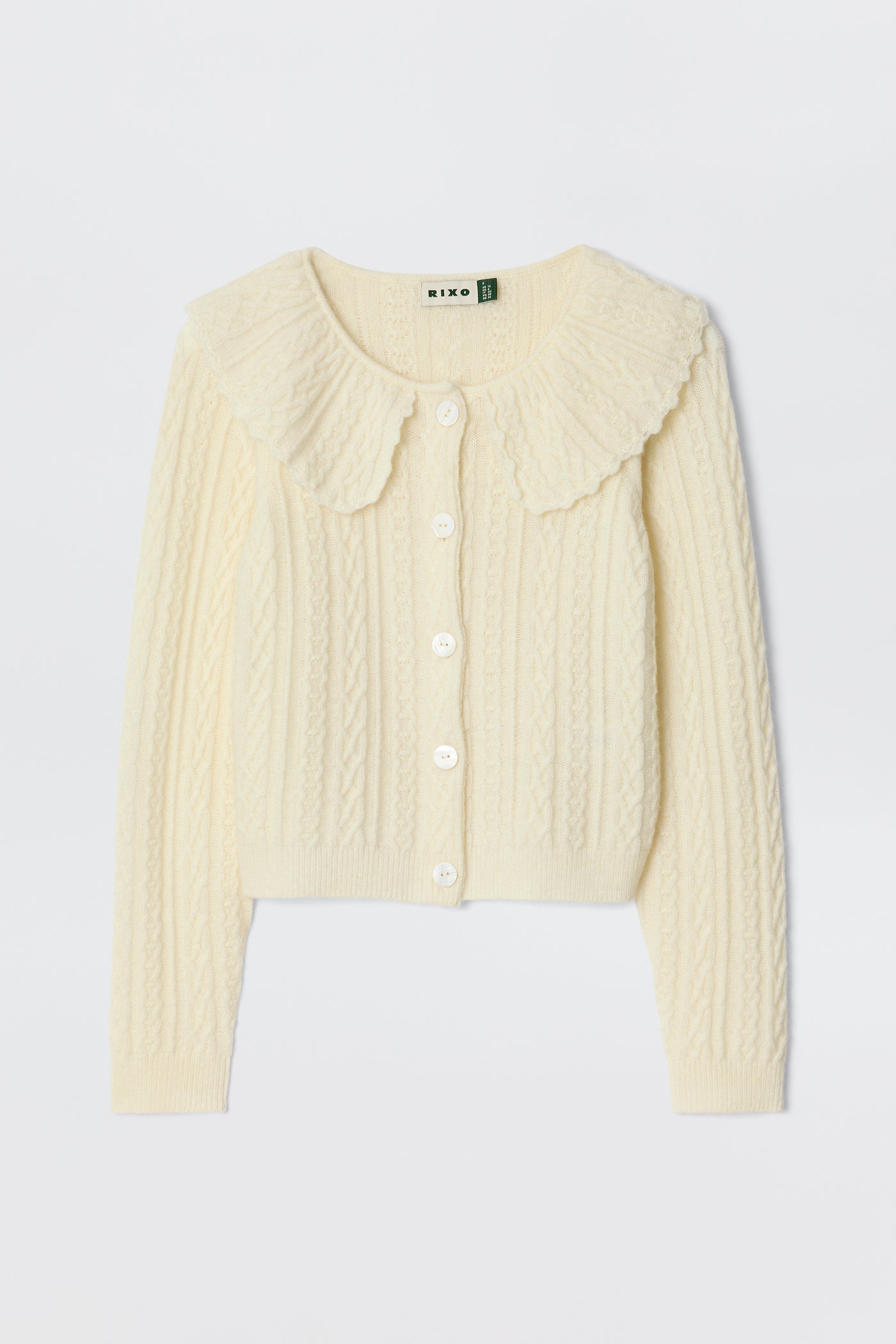 The Jelena - Cream is a cream-colored, long-sleeve cropped cable knit cardigan with white buttons and a large scalloped collar—ideal for layering between seasons. Laid flat on a white background.