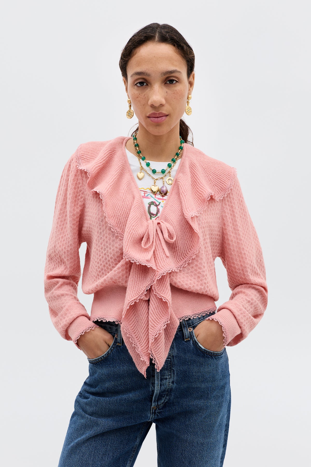 A woman wears the Kianna - Blush pink ruffled transitional knit cardigan over a printed white shirt, blue jeans, gold earrings, and layered necklaces, standing against a plain light gray background facing the camera with hands in her pockets.