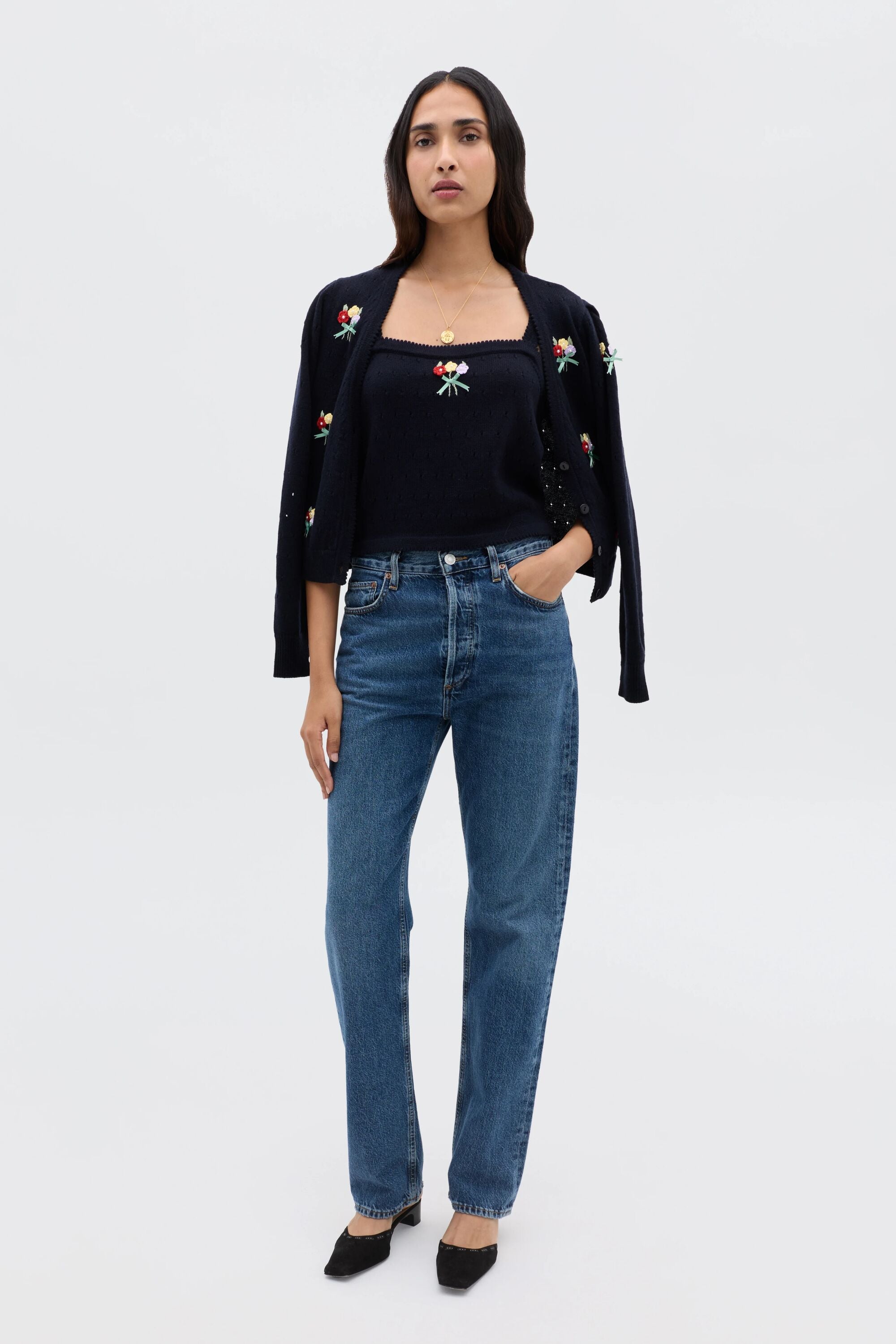 A woman stands facing forward against a plain white background, wearing the Merinda - Floret Embroidery Deep Navy twinset draped over her shoulders, a matching navy top, blue jeans, and black shoes with one hand in her pocket.