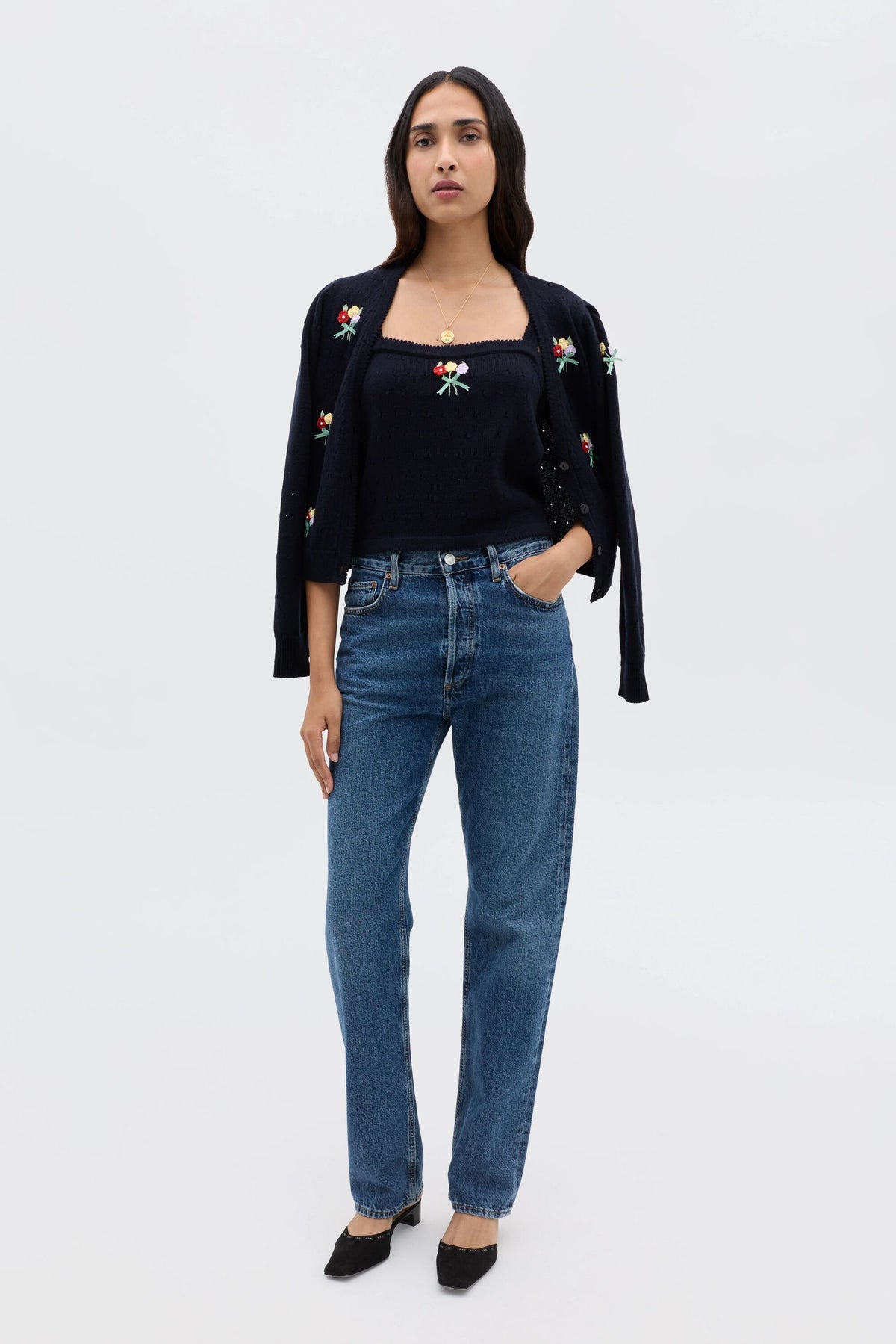A woman stands facing forward against a plain white background, wearing the Merinda - Floret Embroidery Deep Navy twinset draped over her shoulders, a matching navy top, blue jeans, and black shoes with one hand in her pocket.