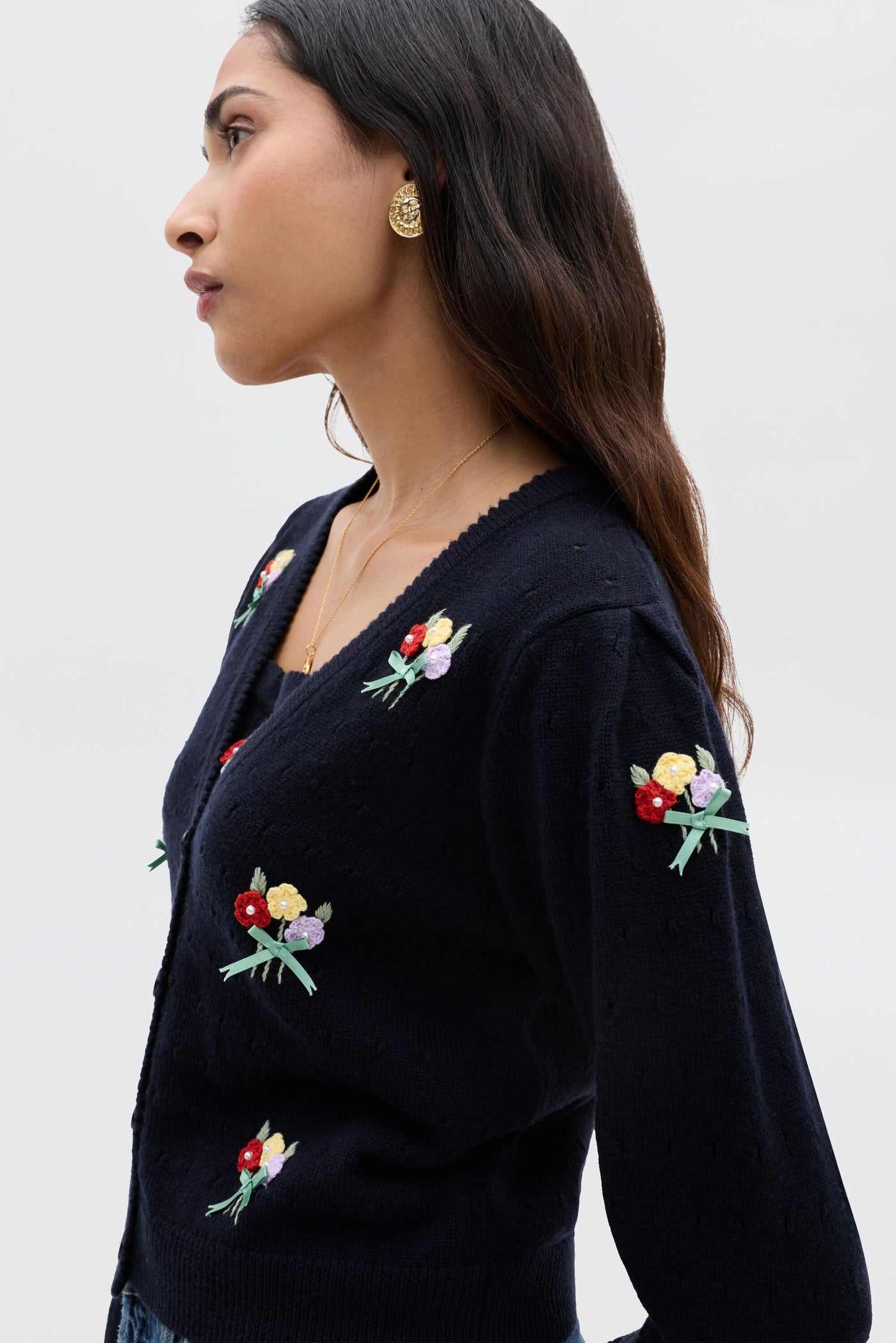 A woman with long dark hair wears the Merinda - Floret Embroidery Deep Navy cardigan, paired with gold earrings and a gold necklace, standing against a plain light background.