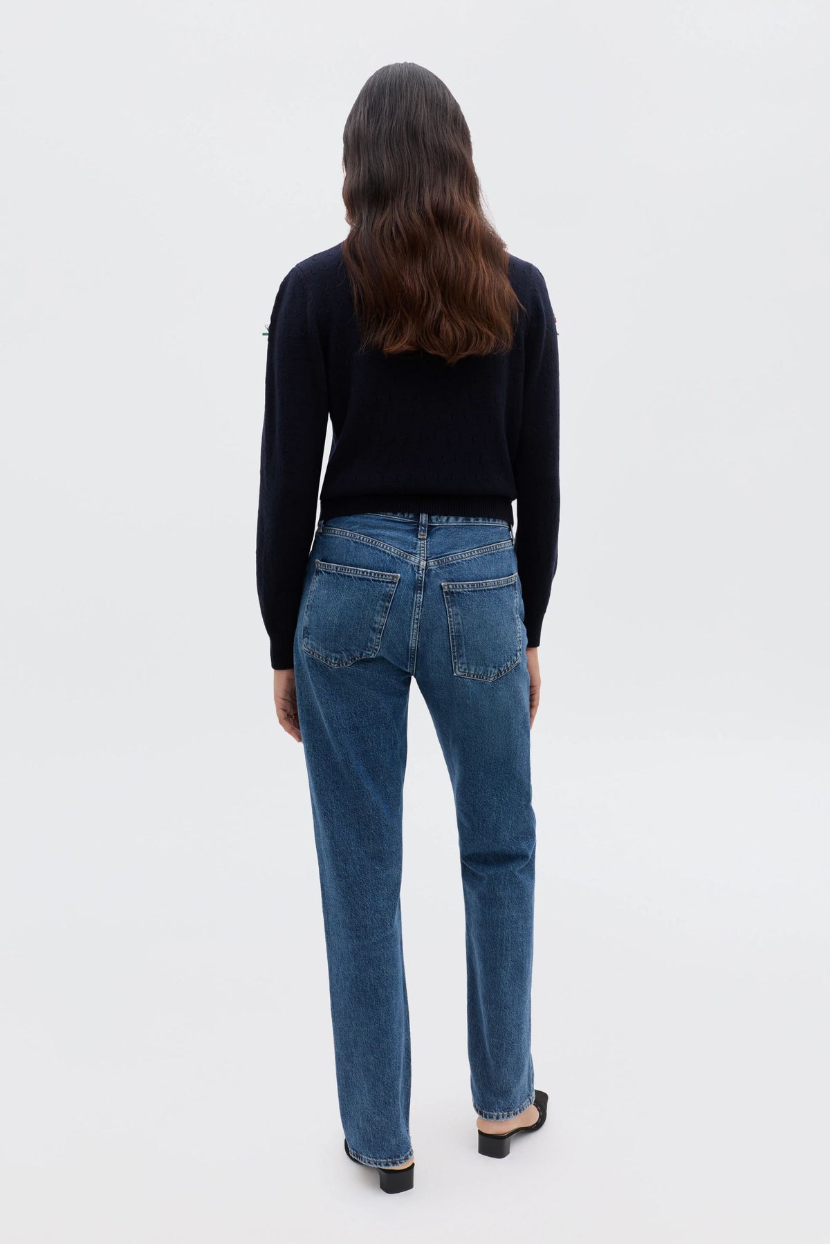 A person with long brown hair, wearing the Merinda - Floret Embroidery Deep Navy sweater and blue jeans, stands facing away from the camera against a plain white background.
