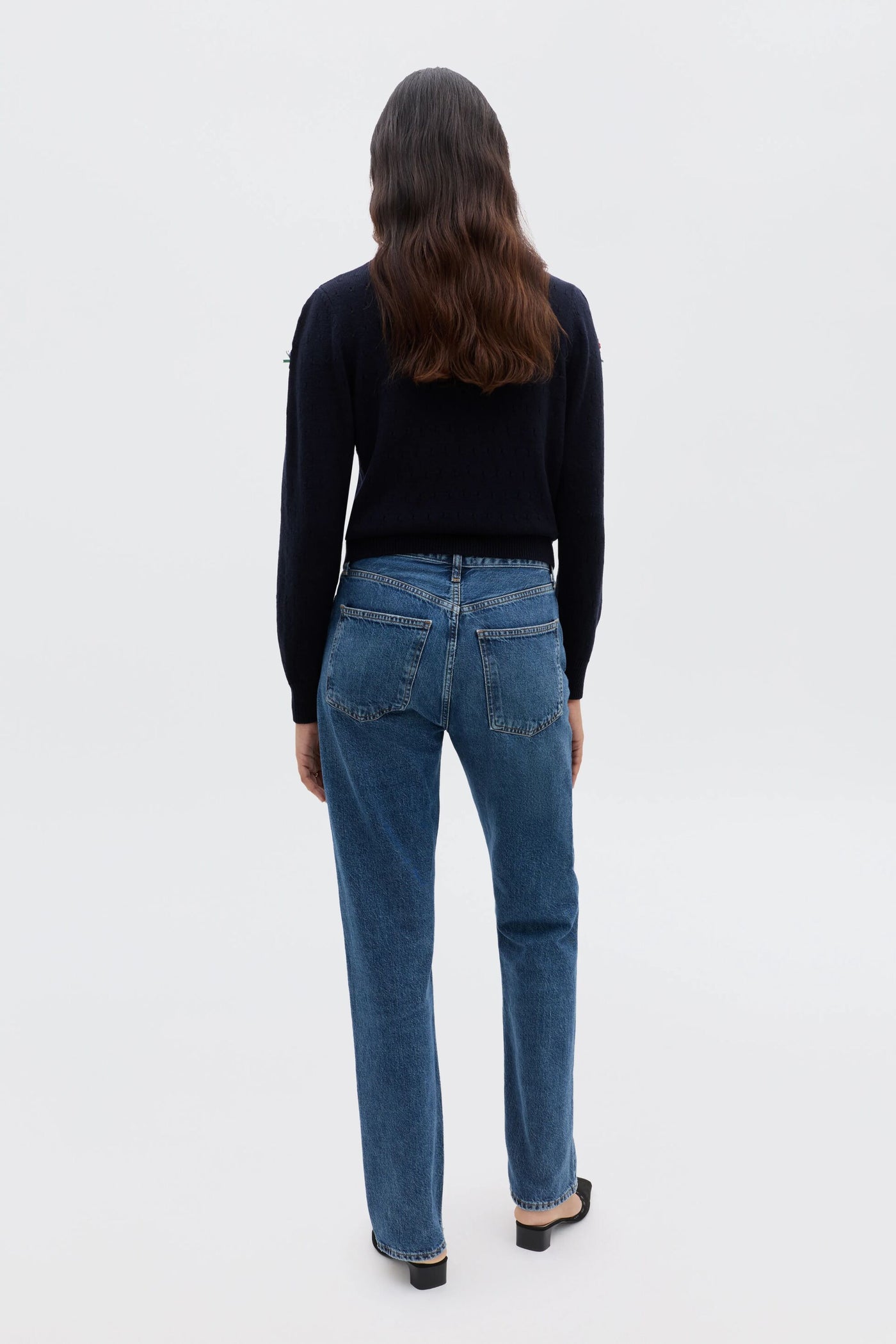 A person with long brown hair, wearing the Merinda - Floret Embroidery Deep Navy sweater and blue jeans, stands facing away from the camera against a plain white background.