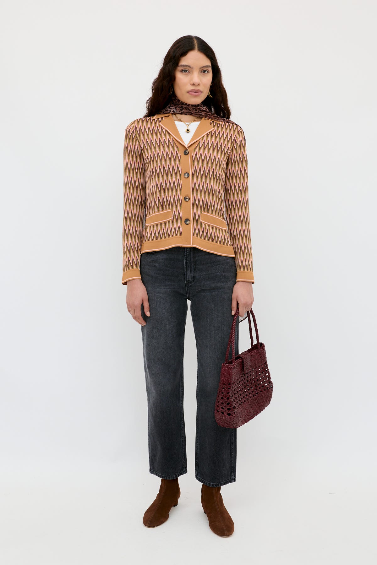 A woman with long dark hair and a neutral expression stands against a white background wearing the Quinley Maeve Jacquard Multi cardigan, paired with dark jeans, brown ankle boots, and holding a burgundy woven handbag.