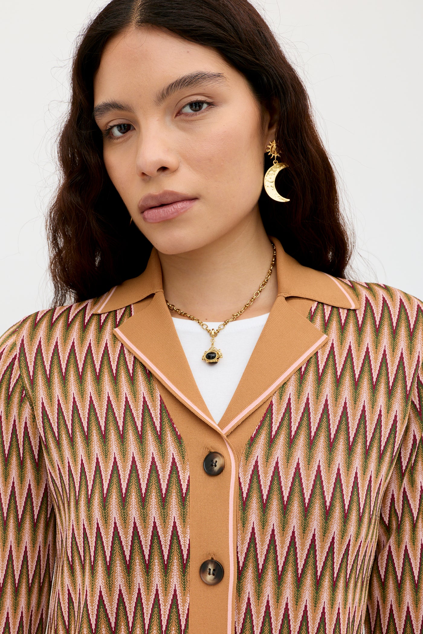 A woman with long wavy hair stands against a plain light background wearing the Quinley - Maeve Jacquard Multi jacket in earth tones, a white top, gold moon earrings, and a gold necklace with a black stone pendant.