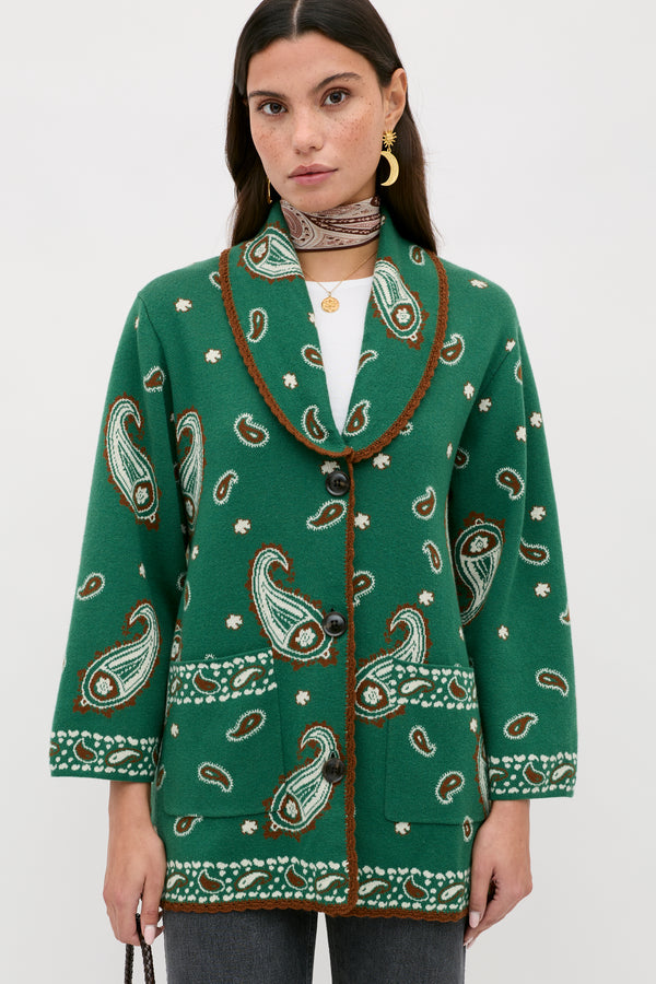 A woman with straight dark hair, a white top, and a patterned neck scarf wears the Morla - Daisy Paisley Green cardigan—featuring a green paisley and floral pattern, brown trim, and large buttons—against a plain background.