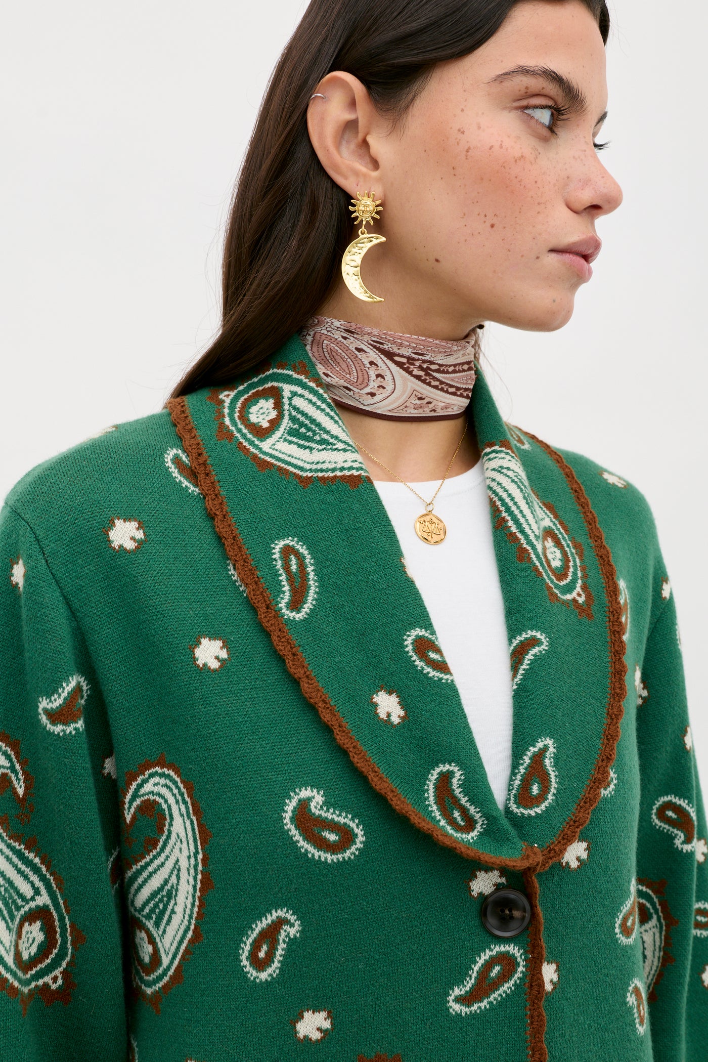 A woman wears the Morla - Daisy Paisley Green cardigan over a white shirt, styled with gold crescent moon and sun earrings, layered gold necklaces, and a patterned neck scarf as she looks to the side.