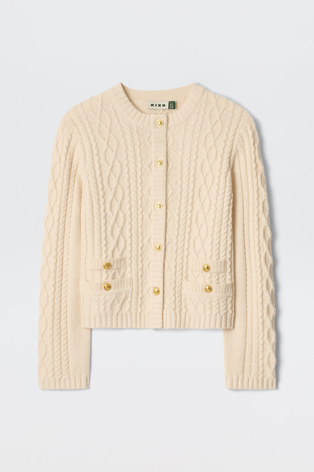 The Kamina - Cream is a cream-colored, long-sleeve cable knit cardigan featuring gold buttons and two front pockets, displayed on a plain white background.