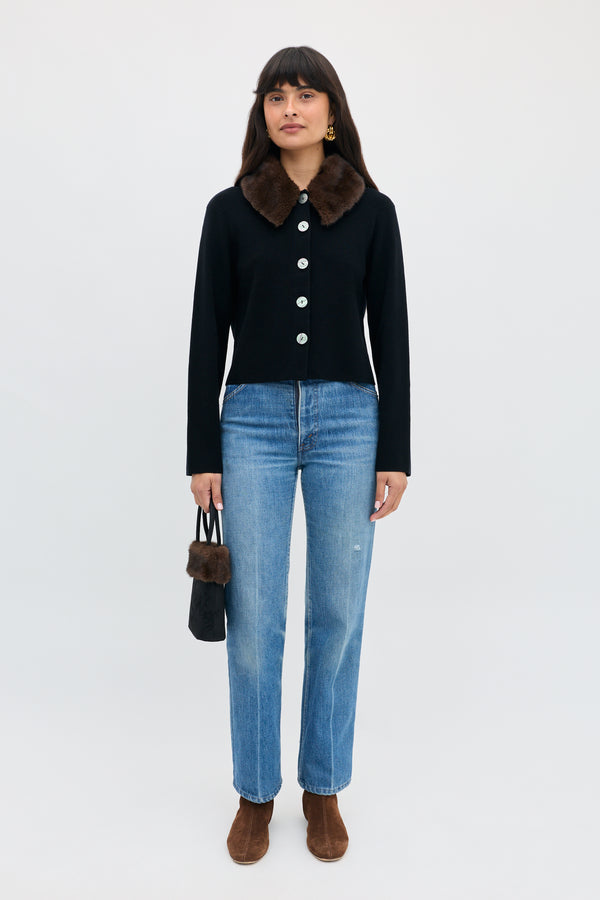 A woman wearing a Jasmin - Black button-down cardigan with a brown faux-fur collar, blue jeans, and brown ankle boots, holding a small black bag with a fur trim.