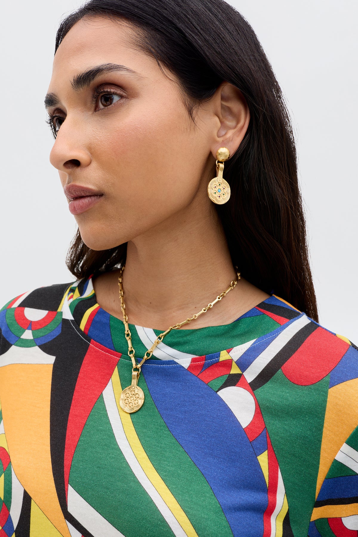 A woman with long dark hair wears the Phaedra - Soho Geo Multi crop top, styled with gold drop earrings and a round pendant necklace, gazing sideways against a plain backdrop.