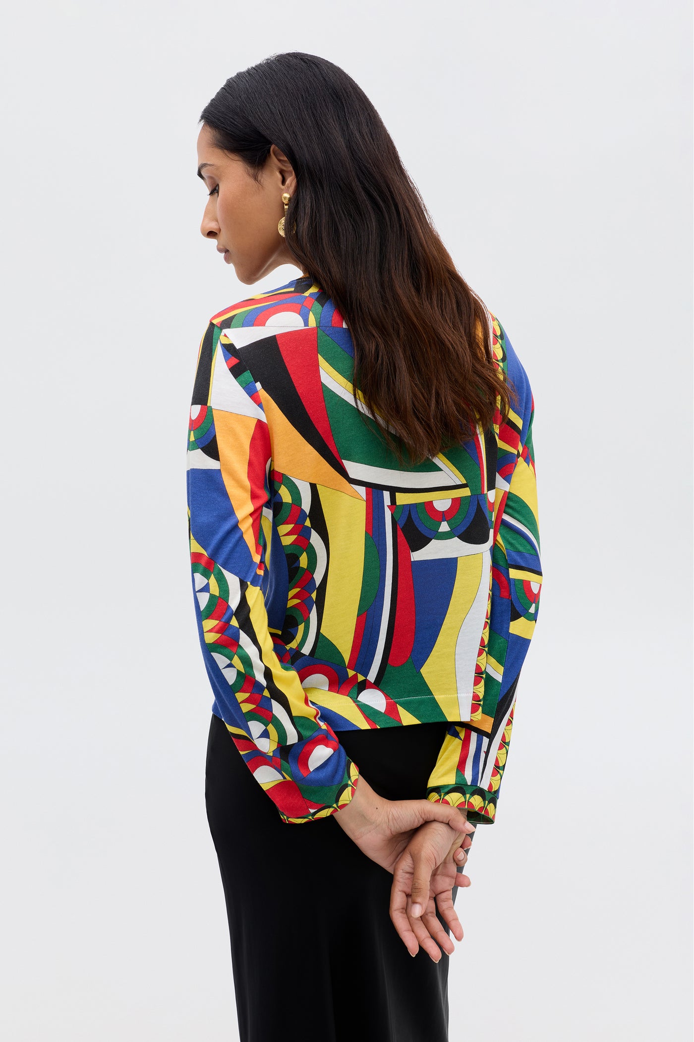 A woman with long, dark hair stands with her back to the camera, hands clasped behind her. She wears the Phaedra crop top in Soho Geo Multi and a black skirt against a plain light background.