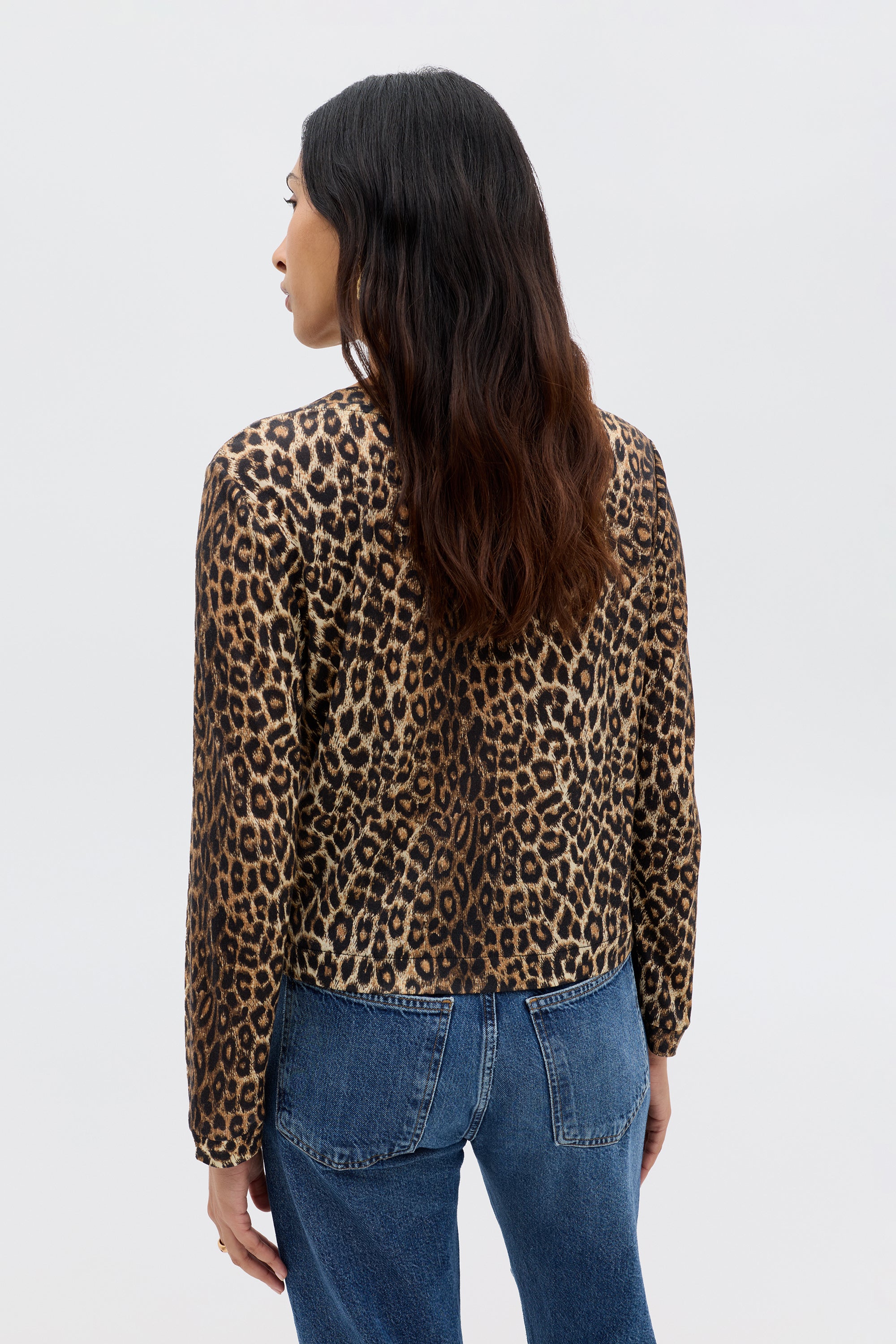 A woman with long, wavy dark hair is wearing the Phaedra - Lyra Leopard Black crop top and blue jeans, standing with her back to the camera against a plain light background.
