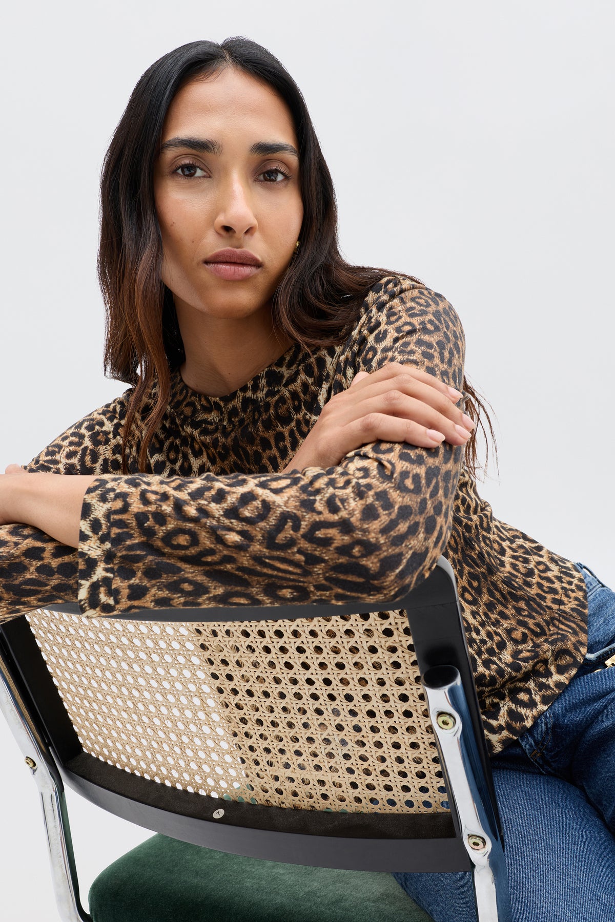 A woman with long dark hair wears the Phaedra - Lyra Leopard Black crop top and blue jeans, leaning on a woven chair and looking confidently at the camera against a plain light background.