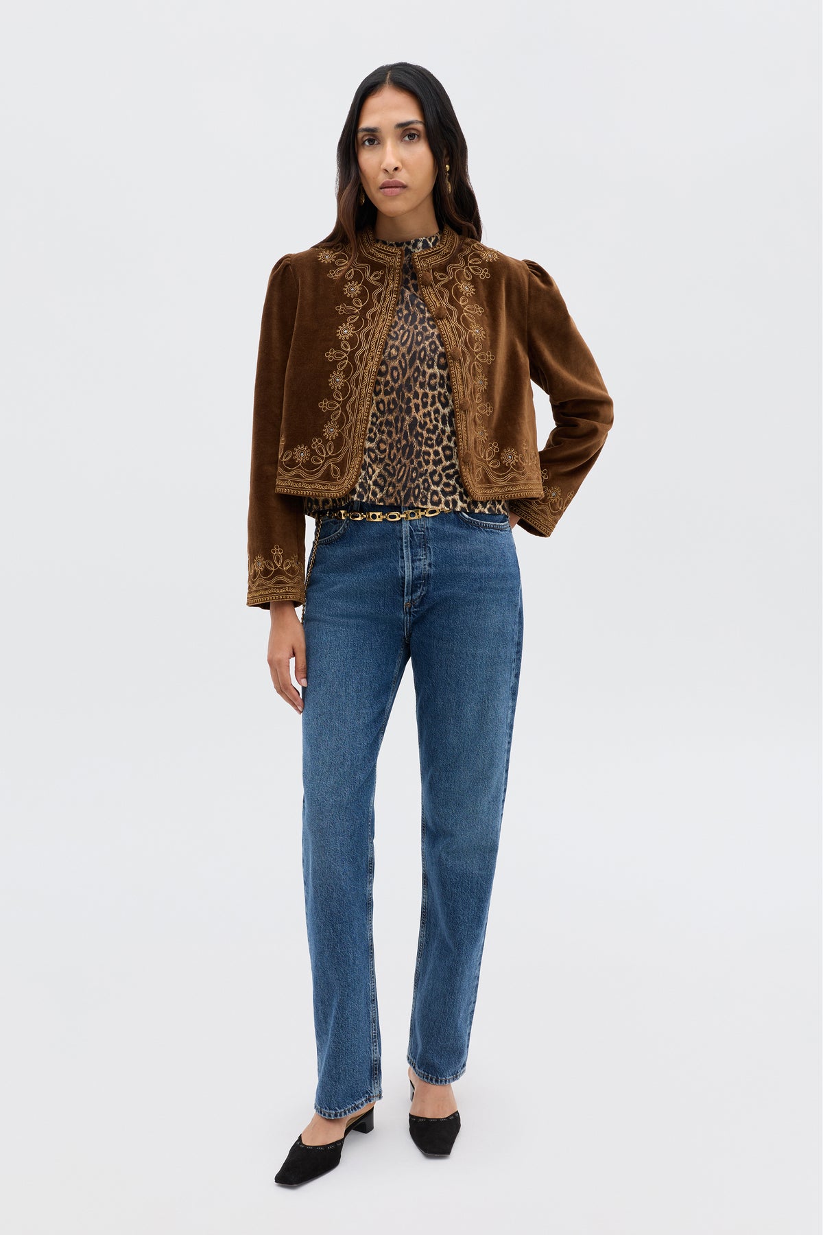A woman with long dark hair and a neutral expression stands against a plain background, wearing a brown embroidered velvet jacket over the Phaedra - Lyra Leopard Black crop top, blue jeans, and black pointed-toe shoes.