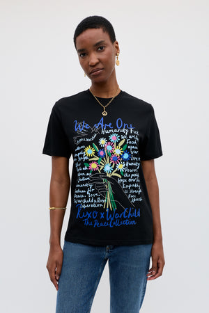 A woman with short hair wears the Charity - Rixo x War Child Black T-shirt with colorful flower graphics and handwritten text. She stands against a plain light background, accessorized with gold jewelry, gazing neutrally at the camera.
