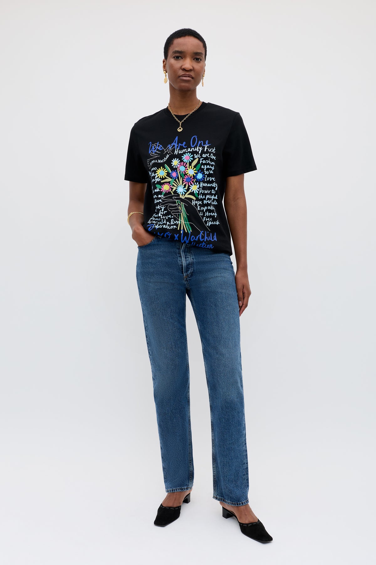 A person faces the camera against a plain background wearing the Charity - Rixo x War Child Black t-shirt with colorful 60s-inspired designs, paired with blue jeans, black heels, and gold jewelry. One hand rests in their pocket.