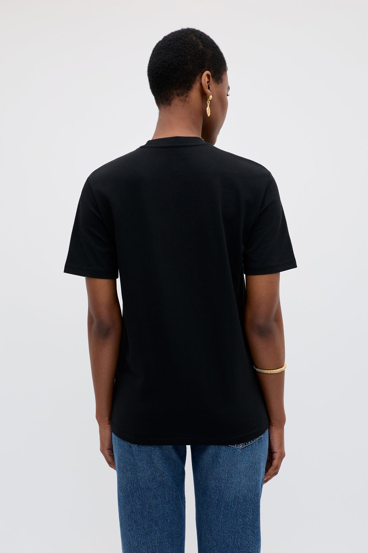 A person with short hair, seen from behind, wears the Charity - Rixo x War Child Black T-shirt, blue jeans, a gold bracelet, and a gold hoop earring against a plain light background.